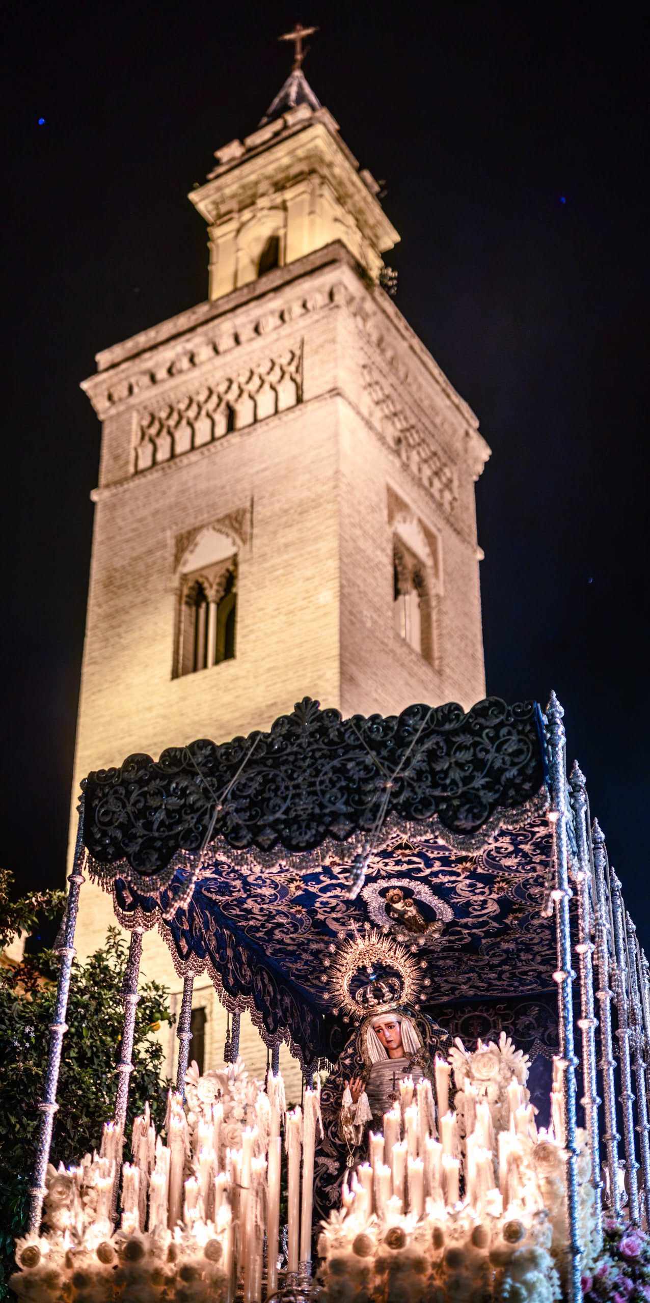 Virgin of Hiniesta's palio float, illuminated by candles, parades past San Marcos Church tower during Holy Week in Seville, Andalusia, Spain.