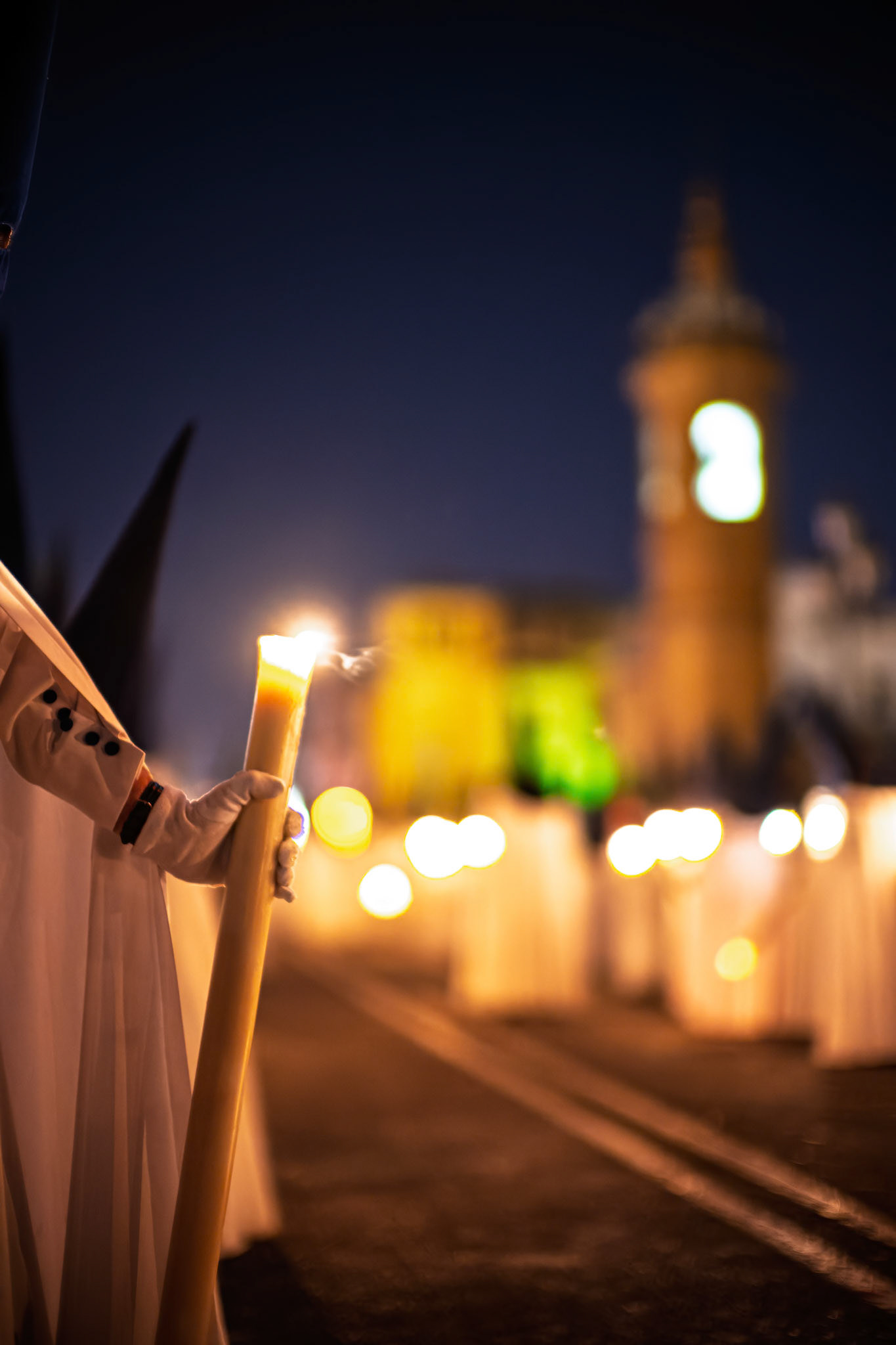 Close-up of a Nazarene from the Estrella brotherhood holding a processional candle on Triana Bridge during Holy Week in Seville. Carmen Chapel is blurred in the background.