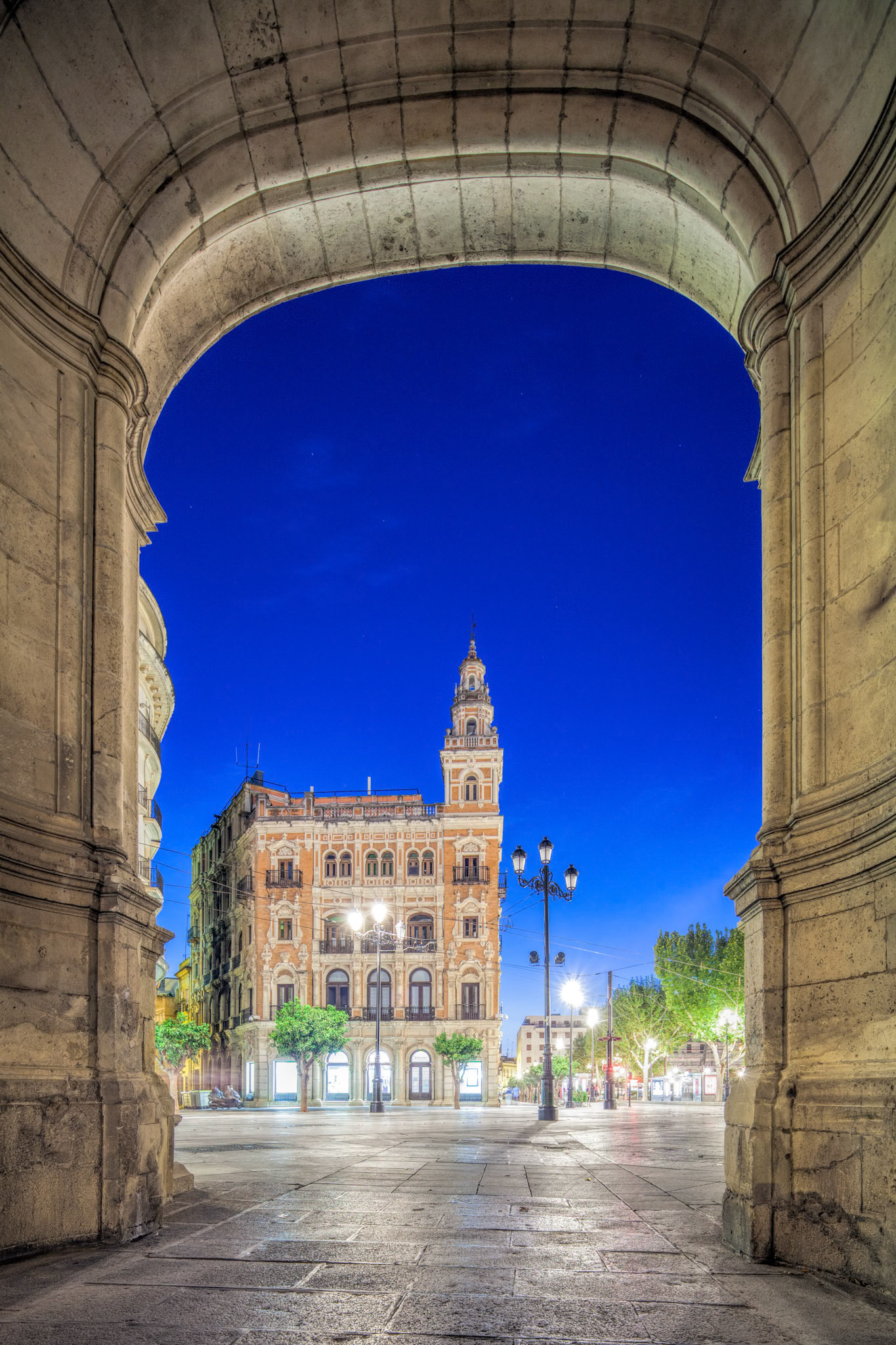 Evening view frames Seville's Telefonica Building through the City Hall's Arquillo, showcasing Spanish architecture with vibrant, dramatic lighting.