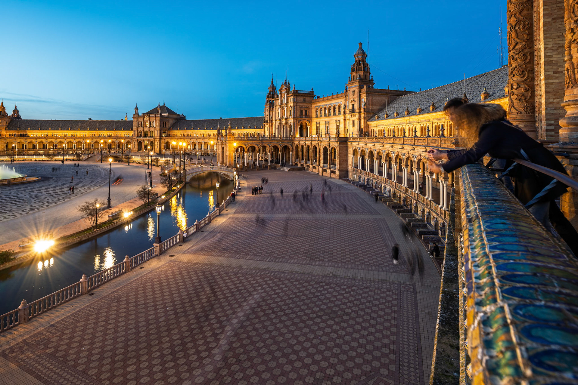 Visitors enjoy a leisurely stroll along the canal of Plaza de España as warm lights illuminate the arches and towers at dusk.