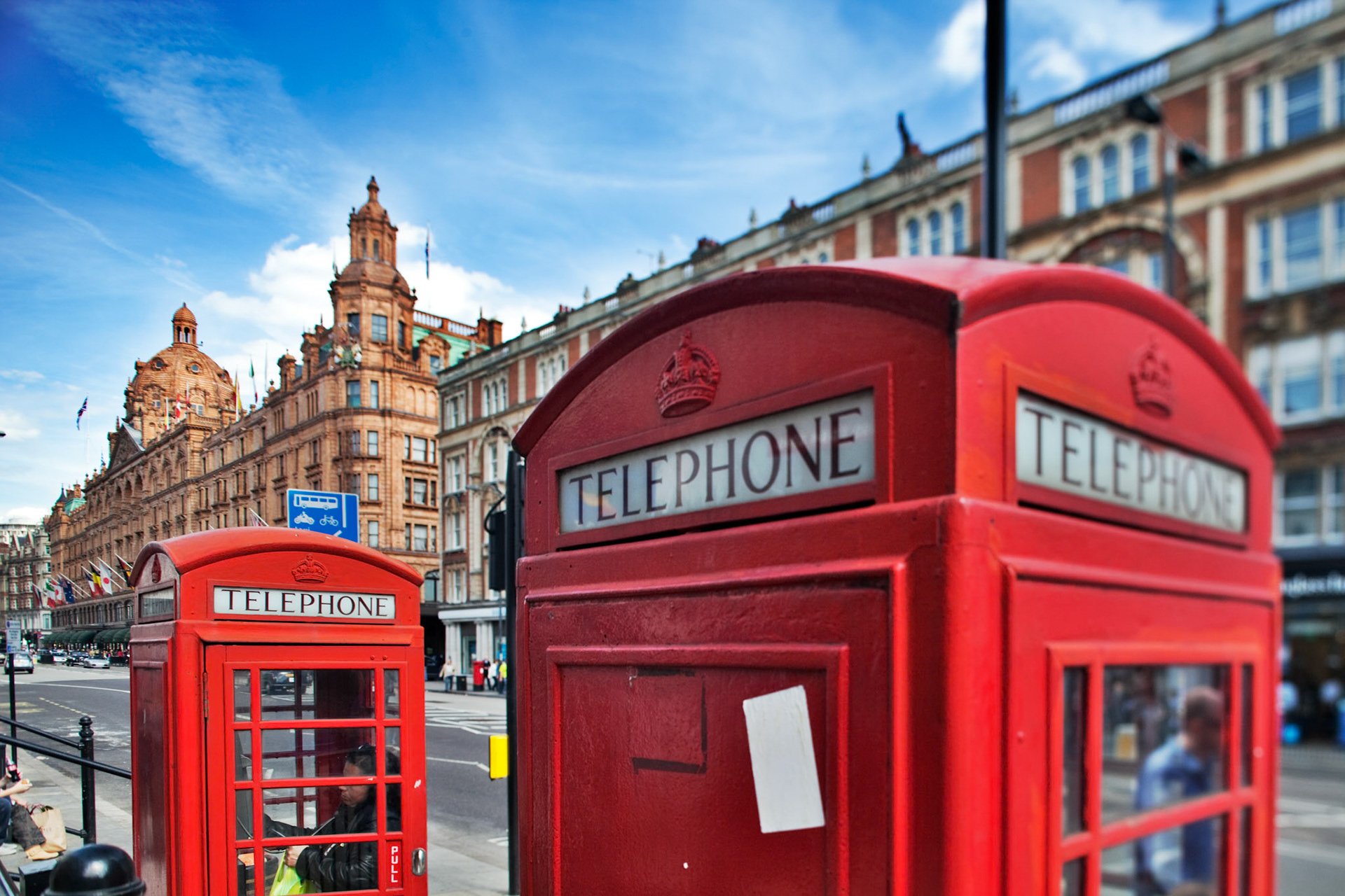 Two iconic red telephone boxes stand on Brompton Road near Harrods, framed by a bright blue sky and bustling city life.