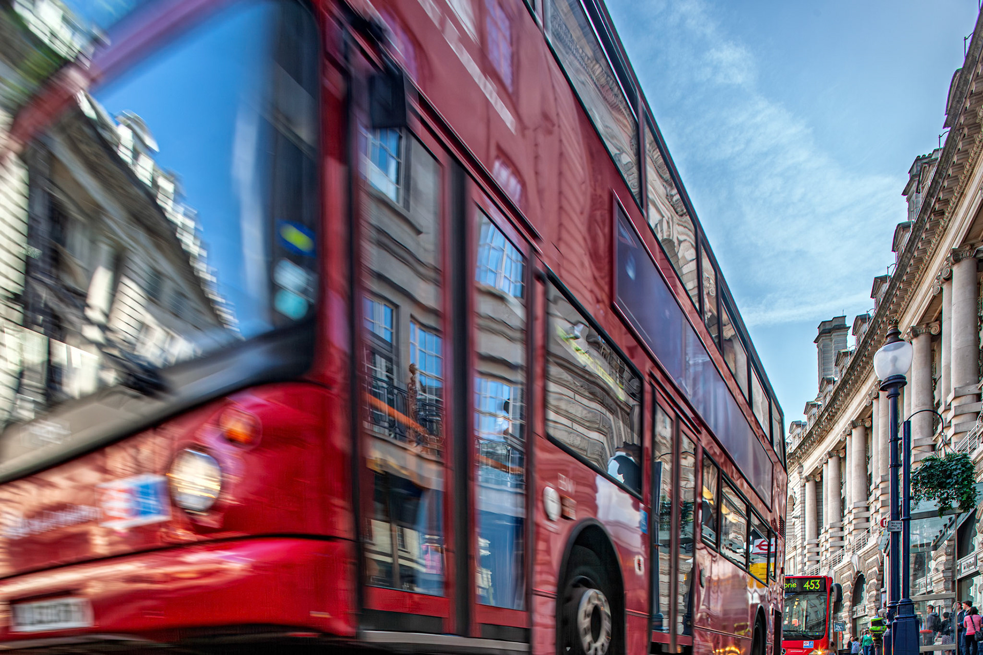 Red double decker buses move along Regent Street in Westminster, London. The scene shows buildings reflecting on the bus windows.