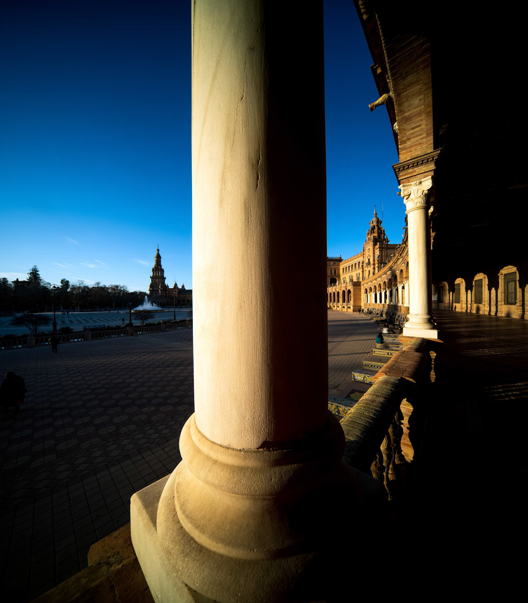 Late afternoon light illuminates the colonnade of Plaza de España in Seville, creating striking silhouettes against the warm stone and tiles.
