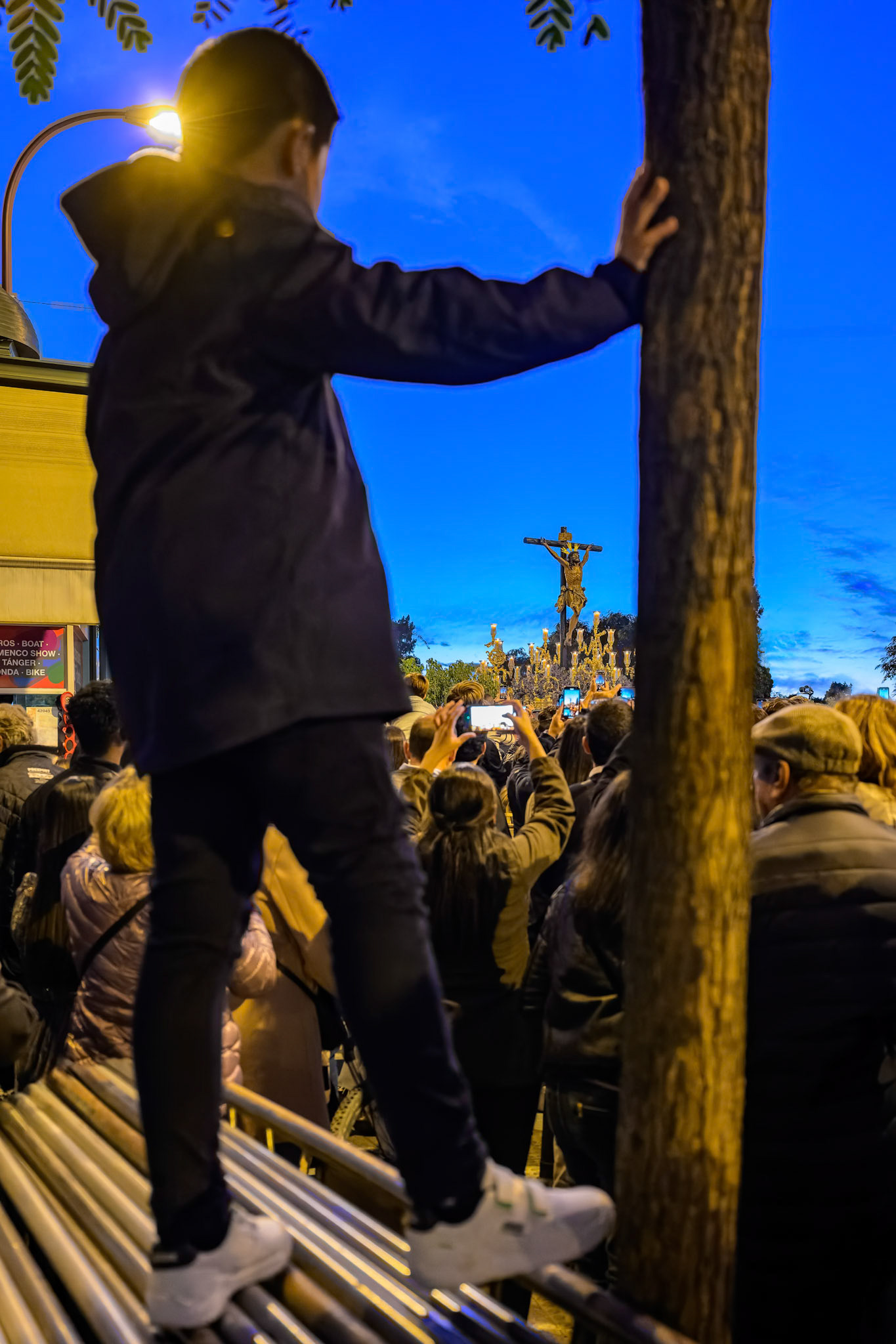 Seville, Spain, Dec 07 2024, In Seville, a child stands on a bench, gazing at the Cristo del Cachorro during a lively procession at dusk.