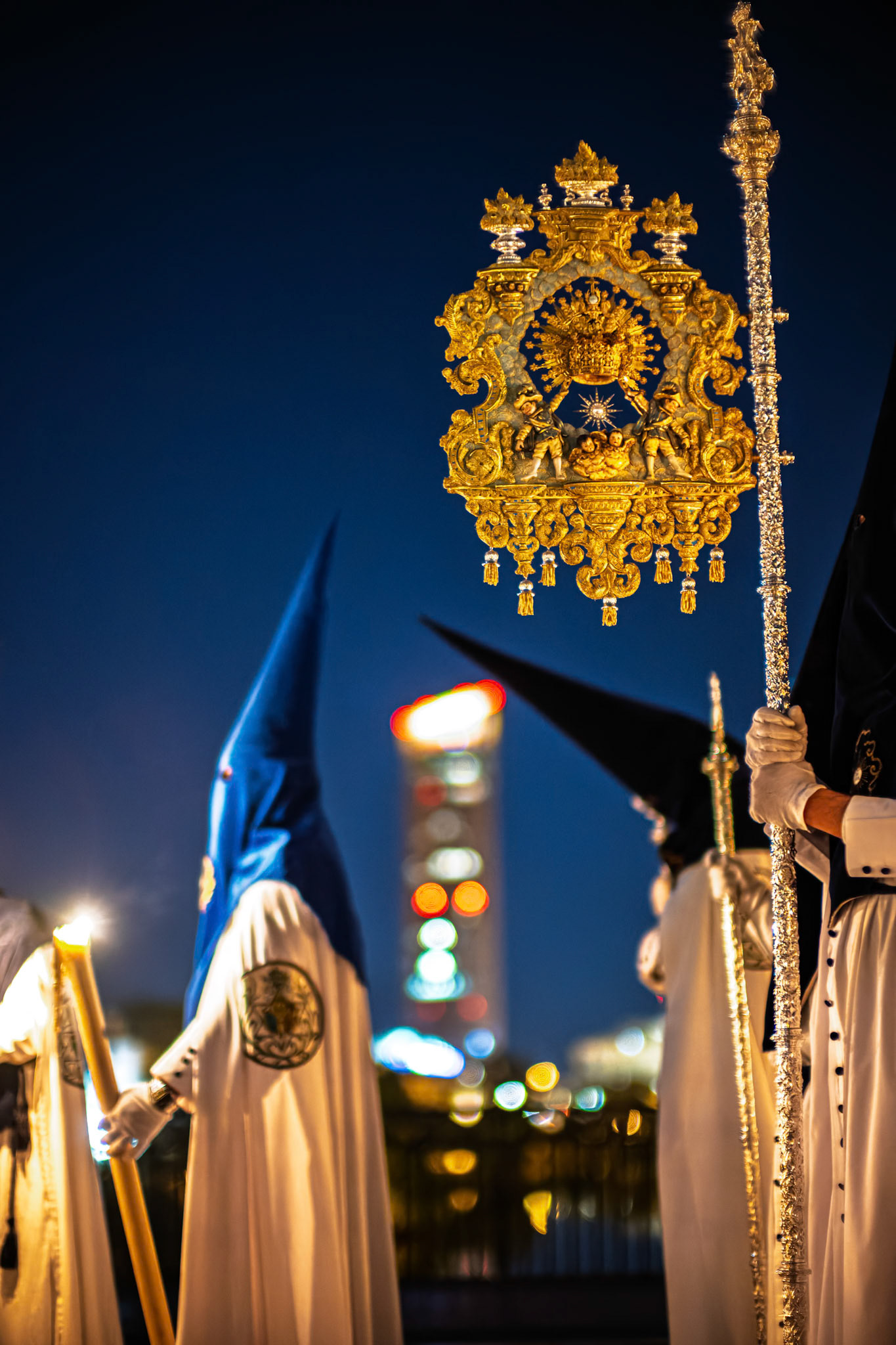 A Nazarene from La Estrella brotherhood holds a banner during a Holy Week procession by night on the Triana Bridge in Seville. Torre Sevilla skyscraper is blurred in the background.