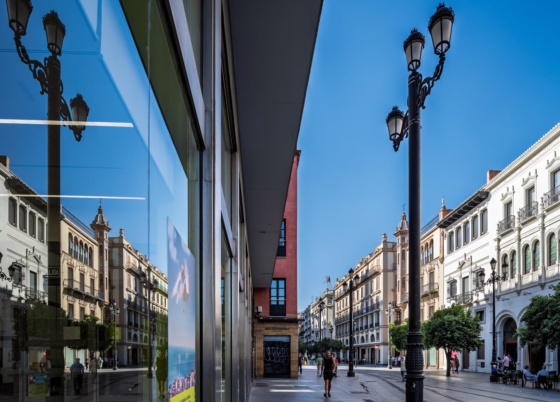 Craftsmanship of early 20th-century buildings shines in reflections on Avenida de la Constitucion, showcasing modern Seville's vibrant atmosphere.