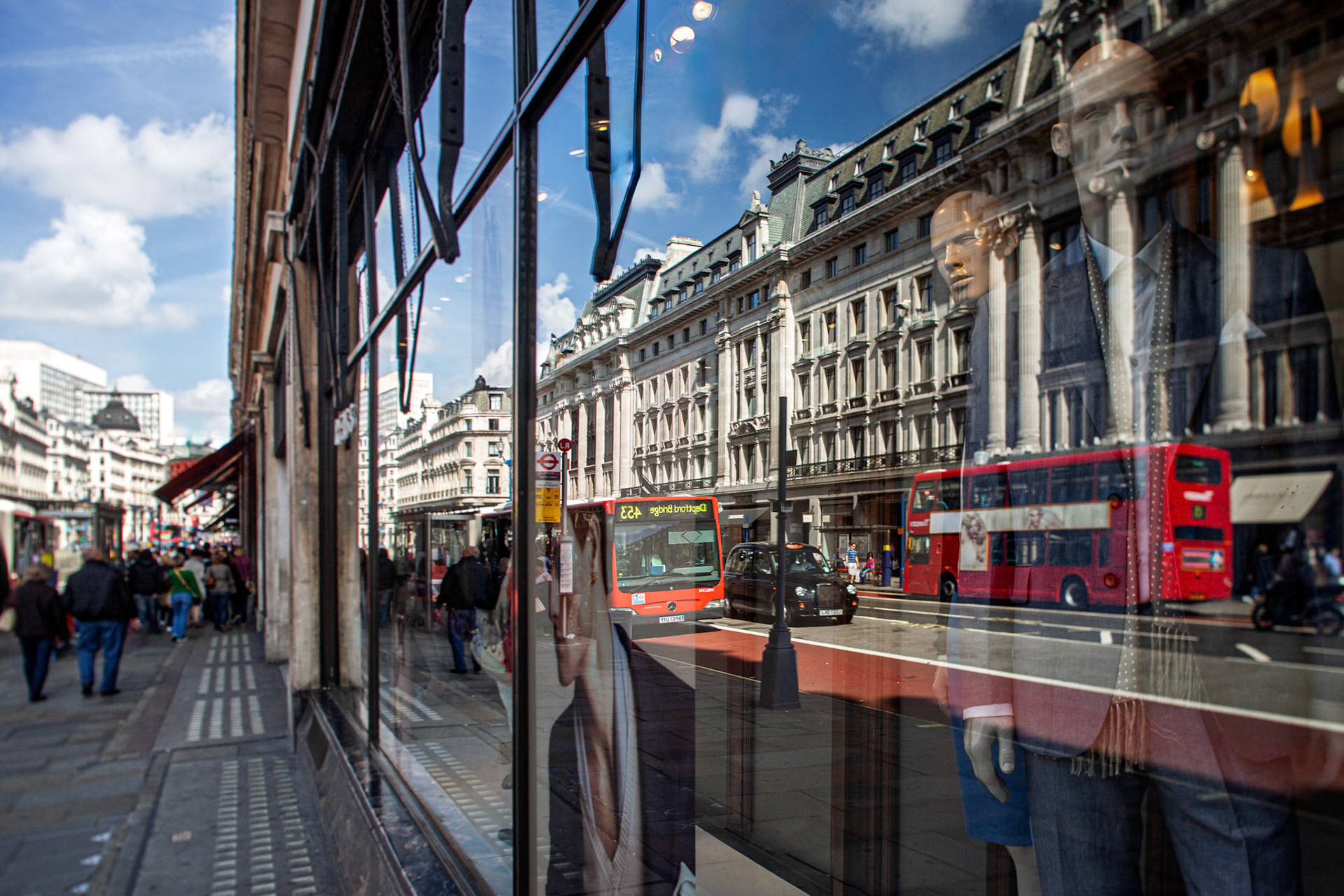 Reflections of shop windows reveal busy streets, double-decker buses, and pedestrians in Westminster, London. A typical day in the city unfolds.
