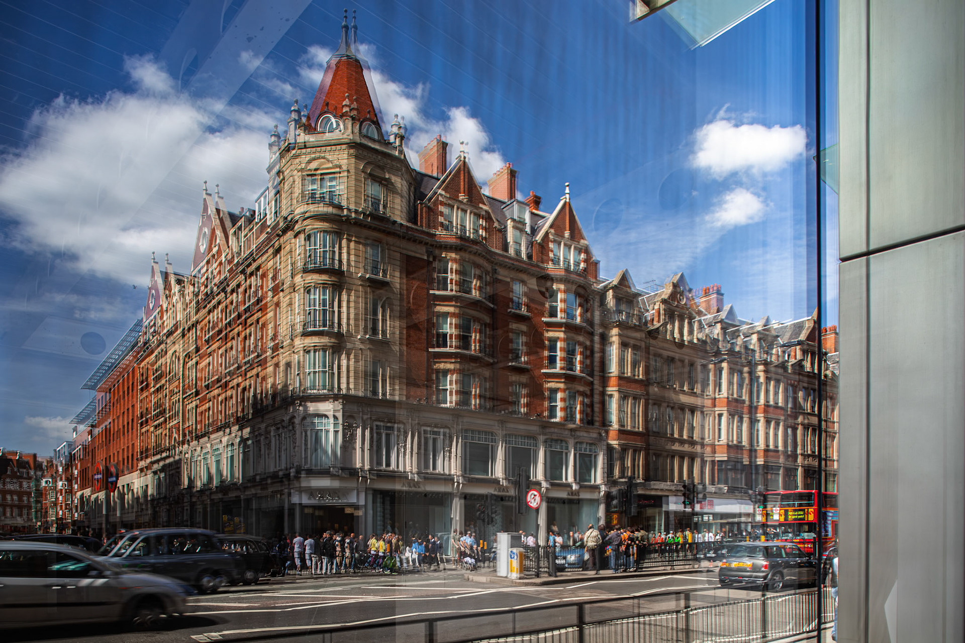 People walk along the street as Knightsbridge Gate reflects on the shop window. The historical building designed in 1902 is a notable sight.
