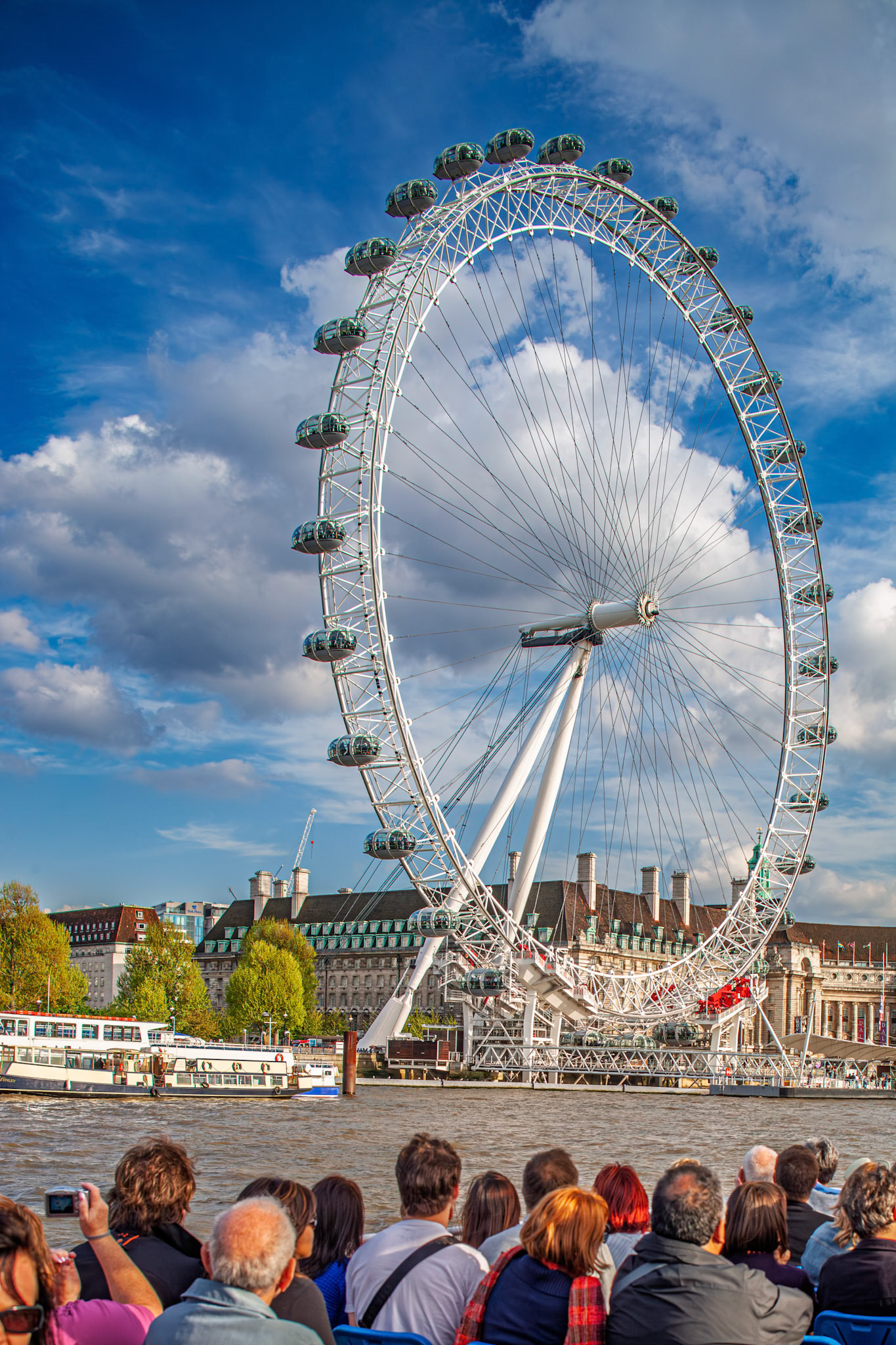 Tourists sit on a boat and look at the London Eye in London. The sky is blue with some clouds, and nearby buildings are visible.