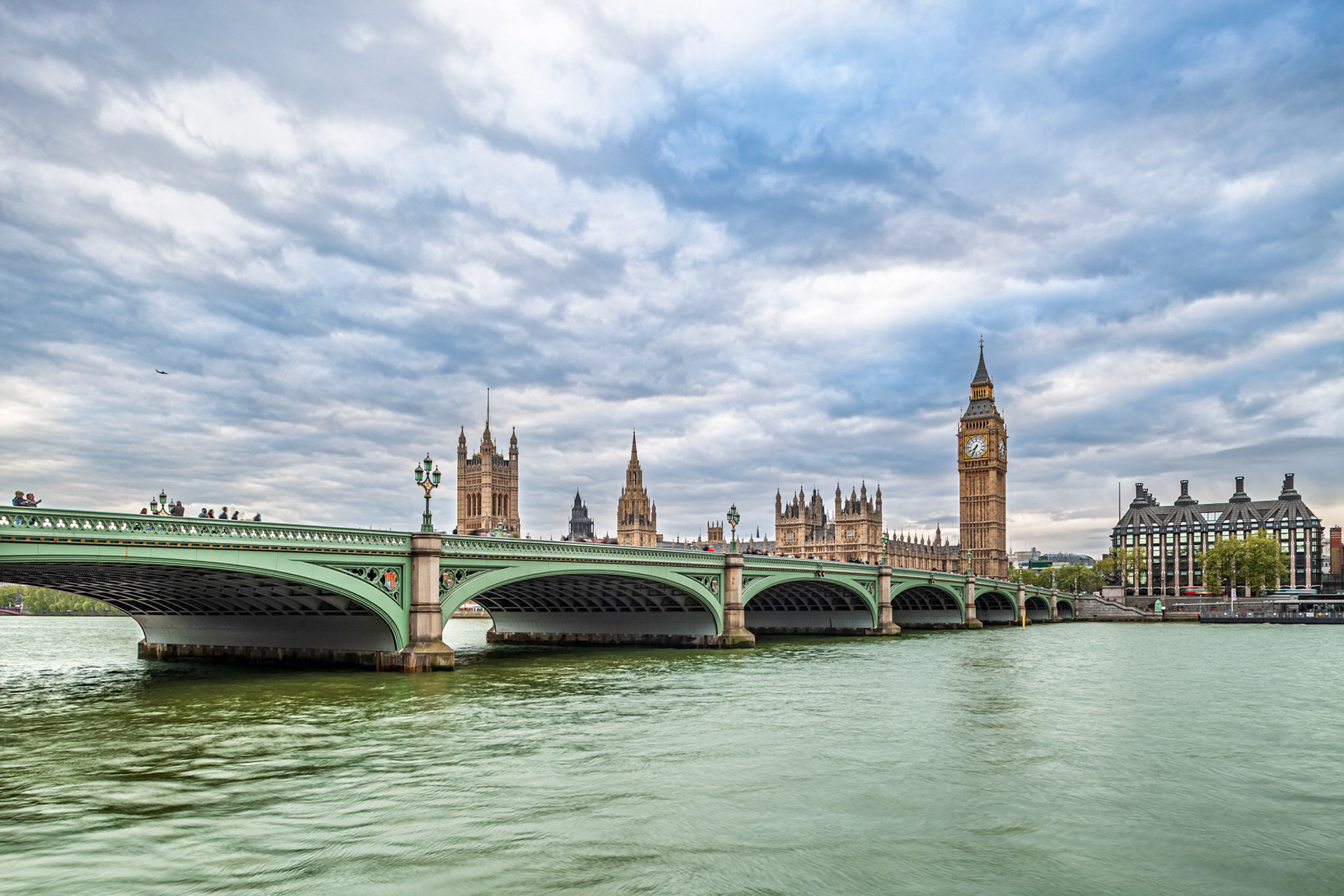 Parliament and Big Ben stand above the River Thames viewed from Westminster Bridge under a cloudy sky in London, UK.