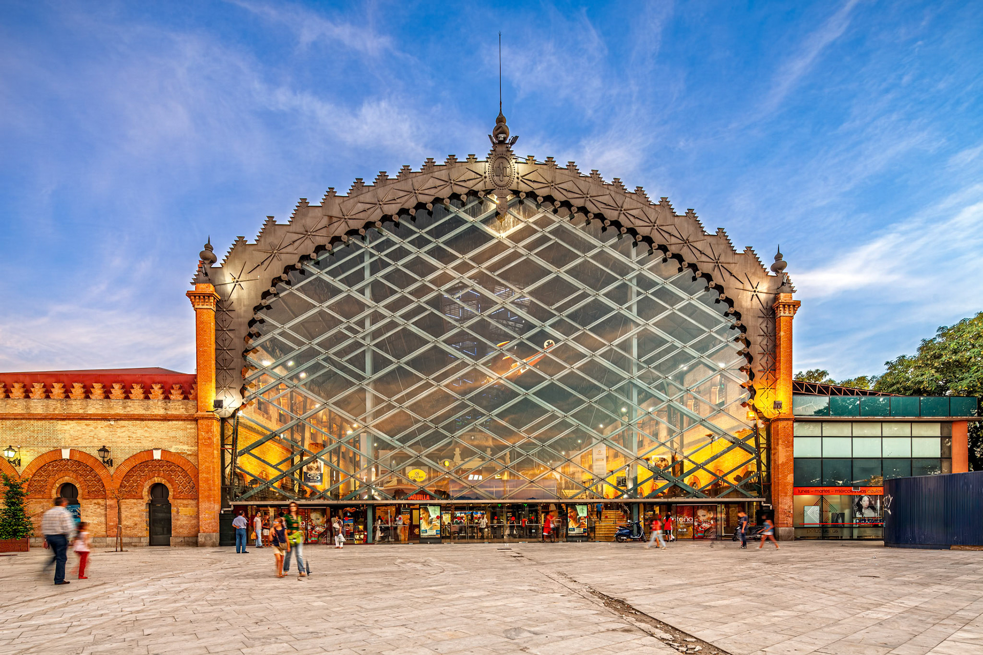 Visitors explore Plaza de Armas, a restored railroad station turned shopping center in Seville, Spain. Exterior view showcases its unique architecture.