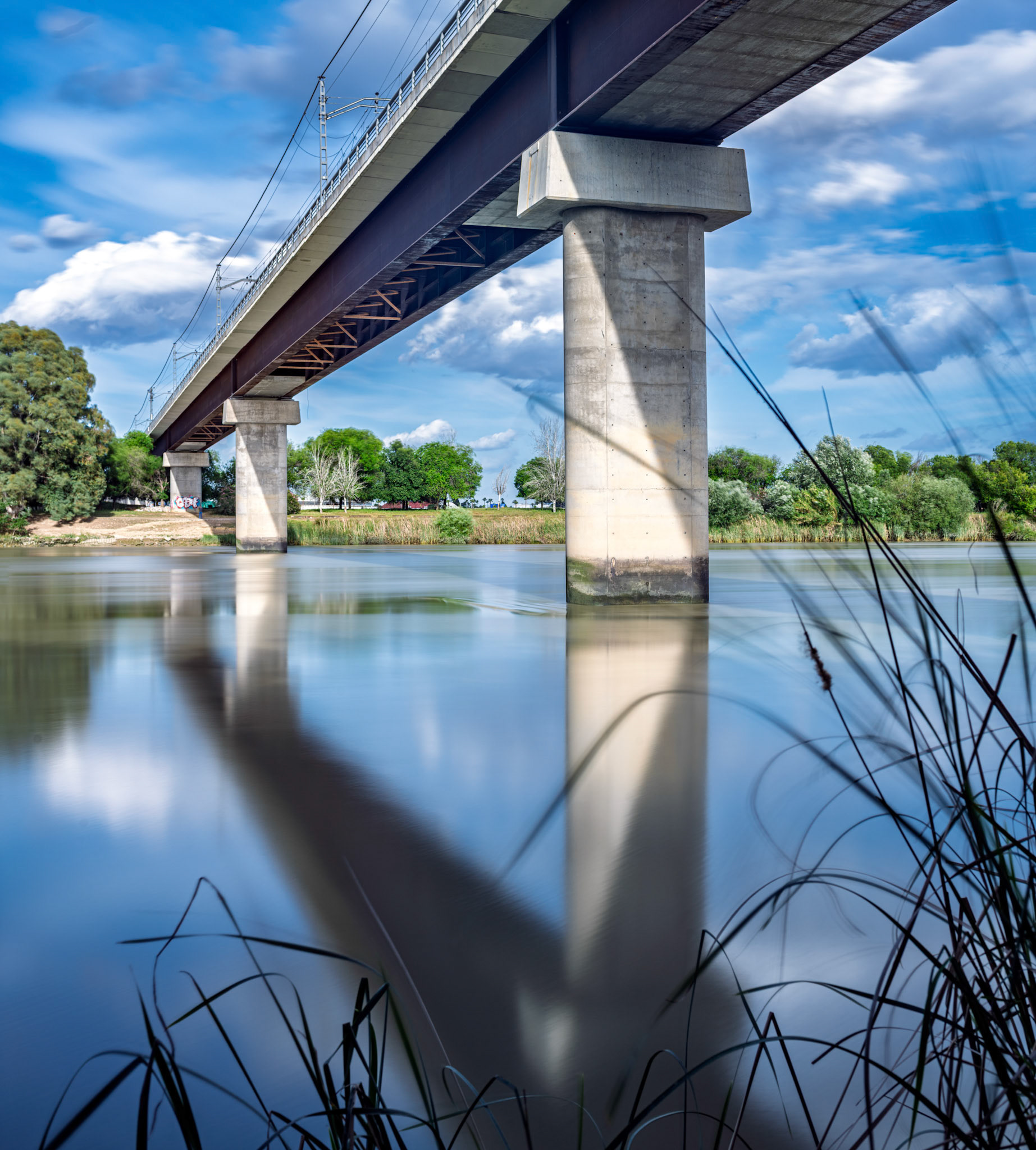 Metro bridge in San Juan de Aznalfarache — captured with long exposure, where concrete pillars and catenary lines stretch over the calm Guadalquivir, blending motion and stillness in Seville’s southern edge.
