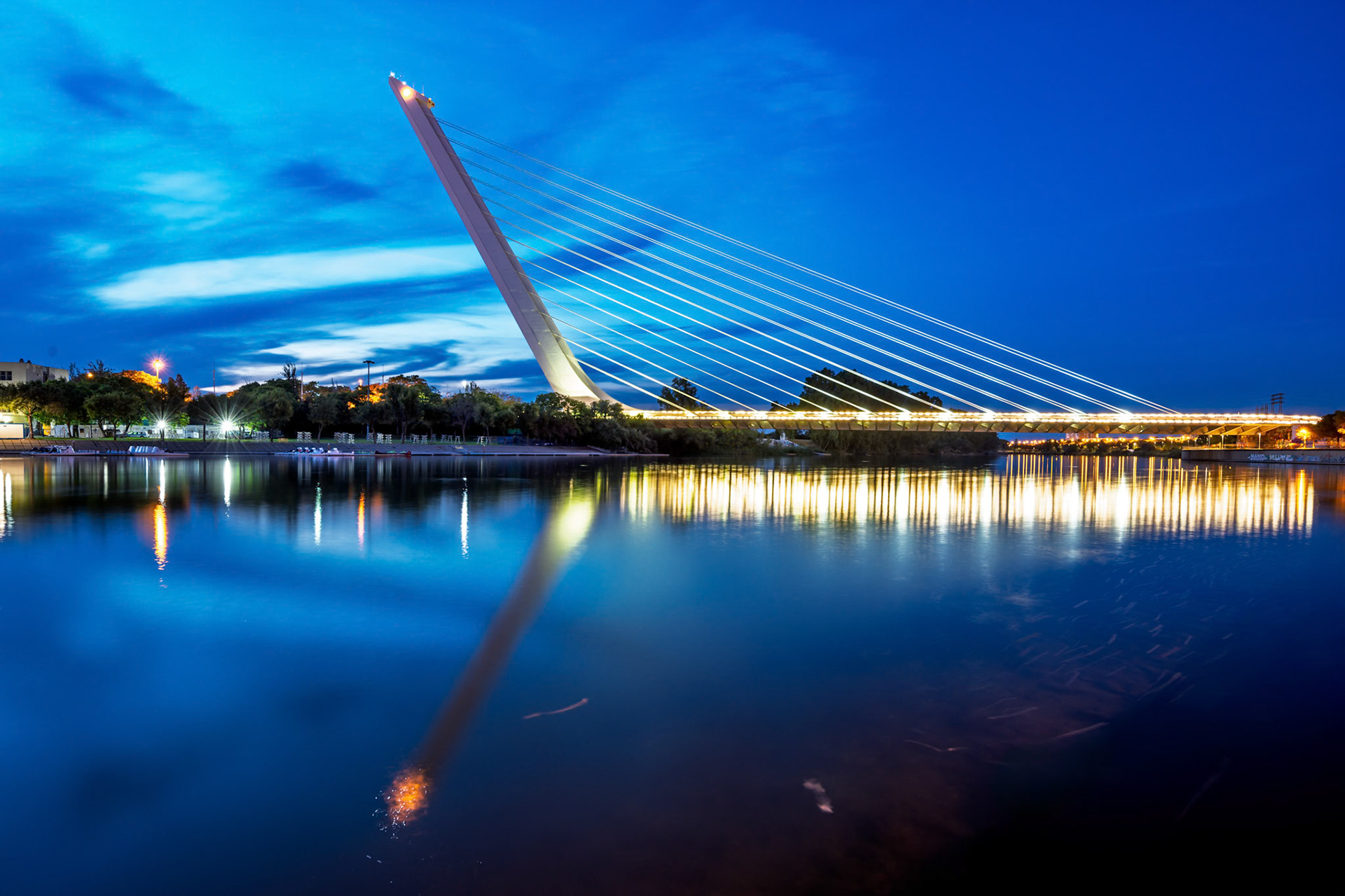Long exposure captures Alamillo Bridge illuminated at dusk, reflecting beautifully in the calm waters of Guadalquivir River in Seville.