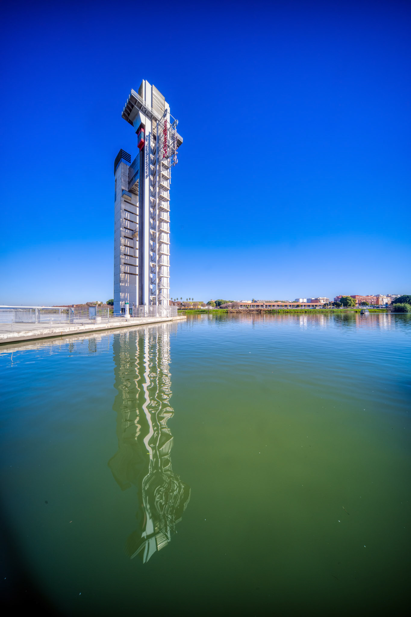Torre Schindler stands tall beside the Guadalquivir River, showcasing its unique design and reflecting beautifully in the calm water.