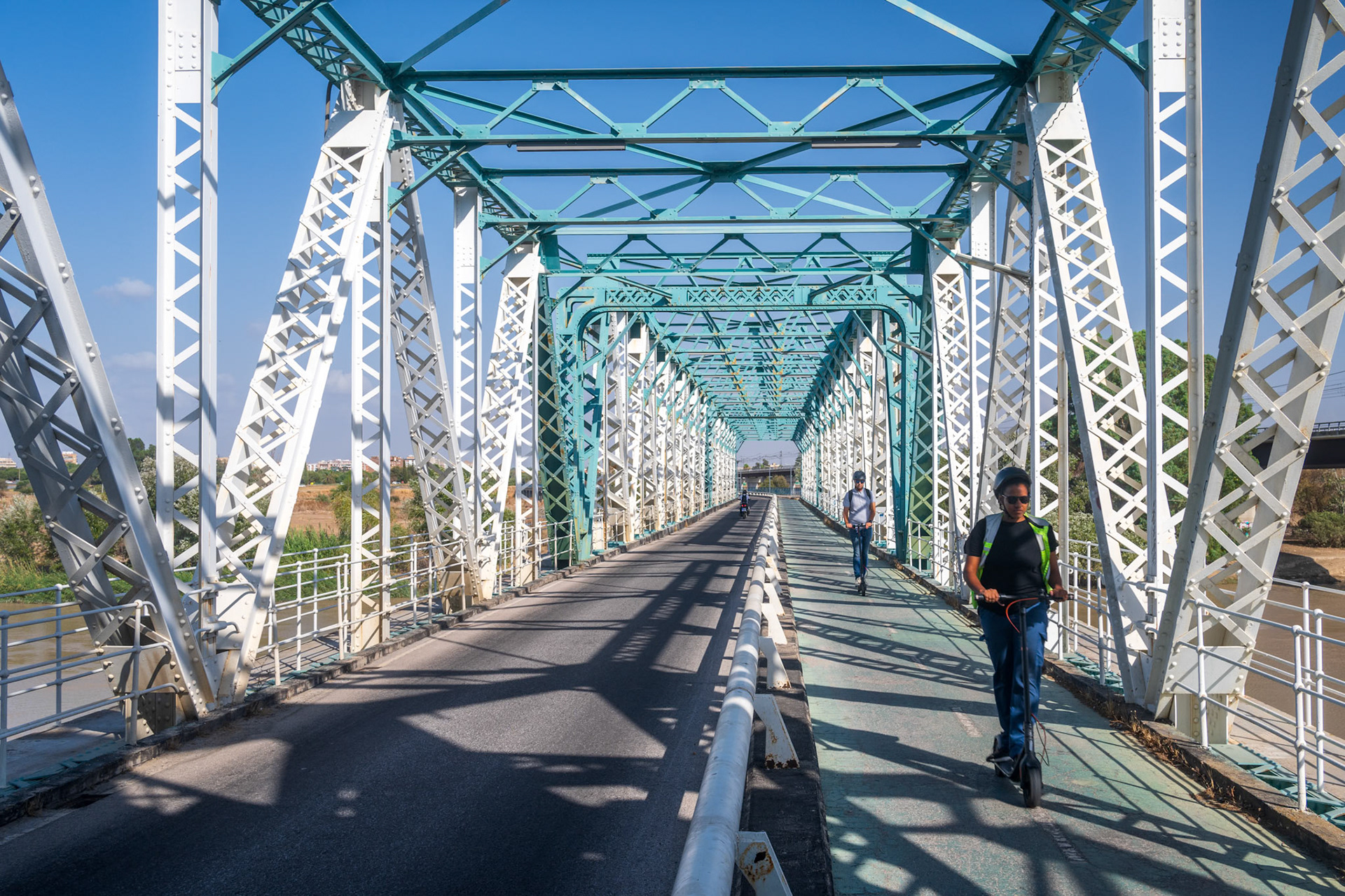 Seville, Spain, Sep 17 2024, Urban scene of people riding electric scooters on the historic San Juan Iron Bridge, built in 1930, over the Guadalquivir River in Seville, Spain. The bridge structure dominates the landscape.