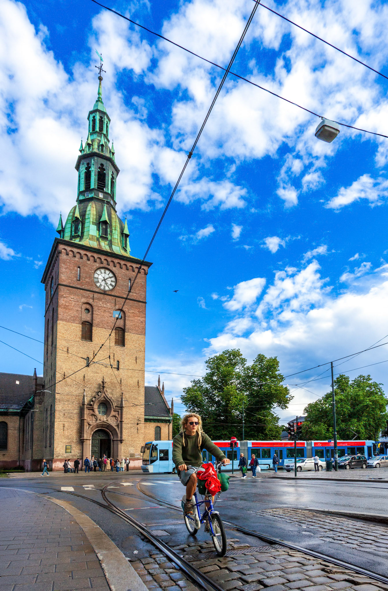 Oslo, Norway, Aug 10 2017, A person cycles by Oslo Cathedral, enjoying the sunny weather, surrounded by vibrant clouds and city life on an active street.