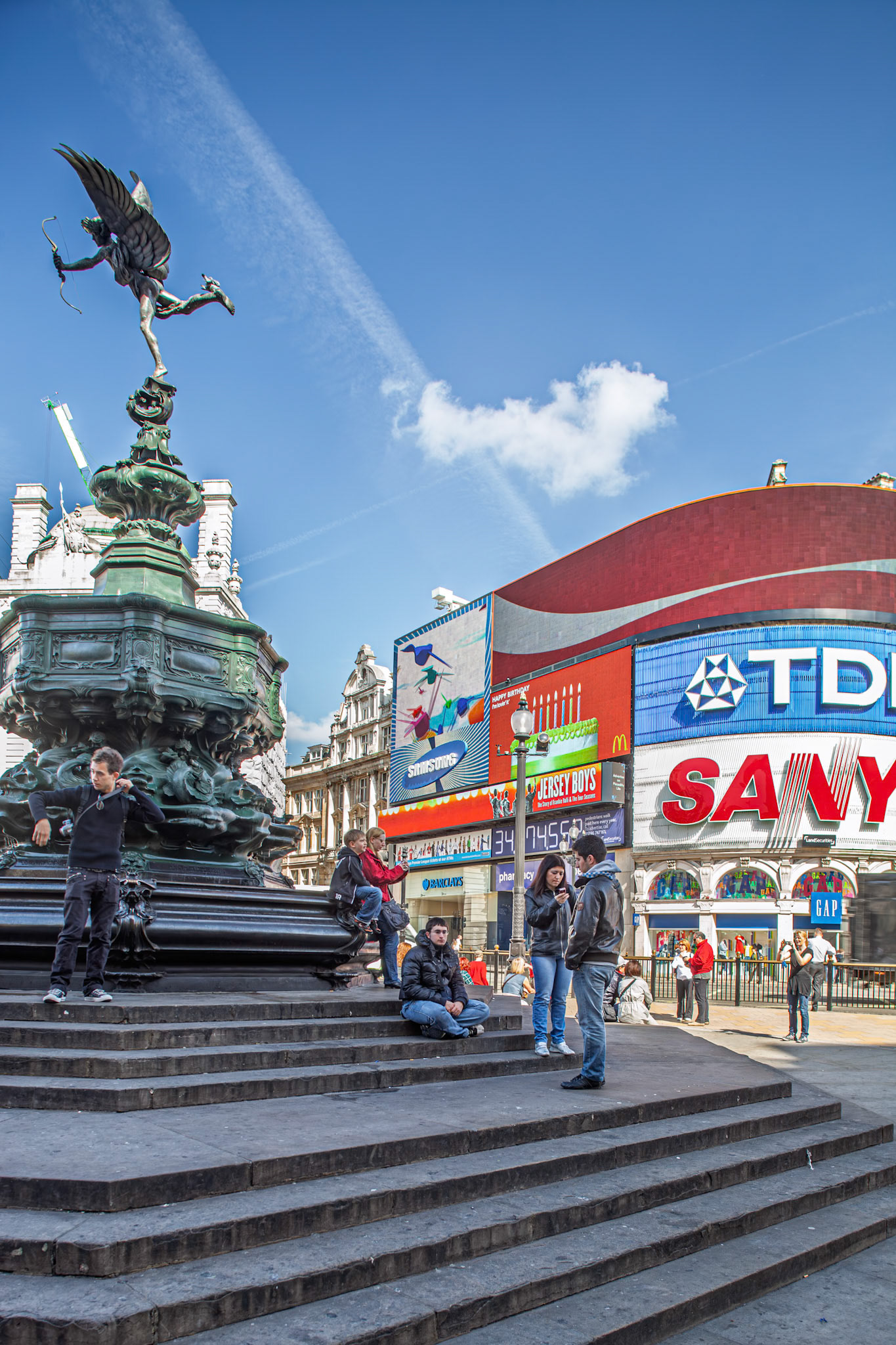 People relax on the steps at Piccadilly Circus in London. Bright signs illuminate the square on a clear day, showing daily activities and advertisements.