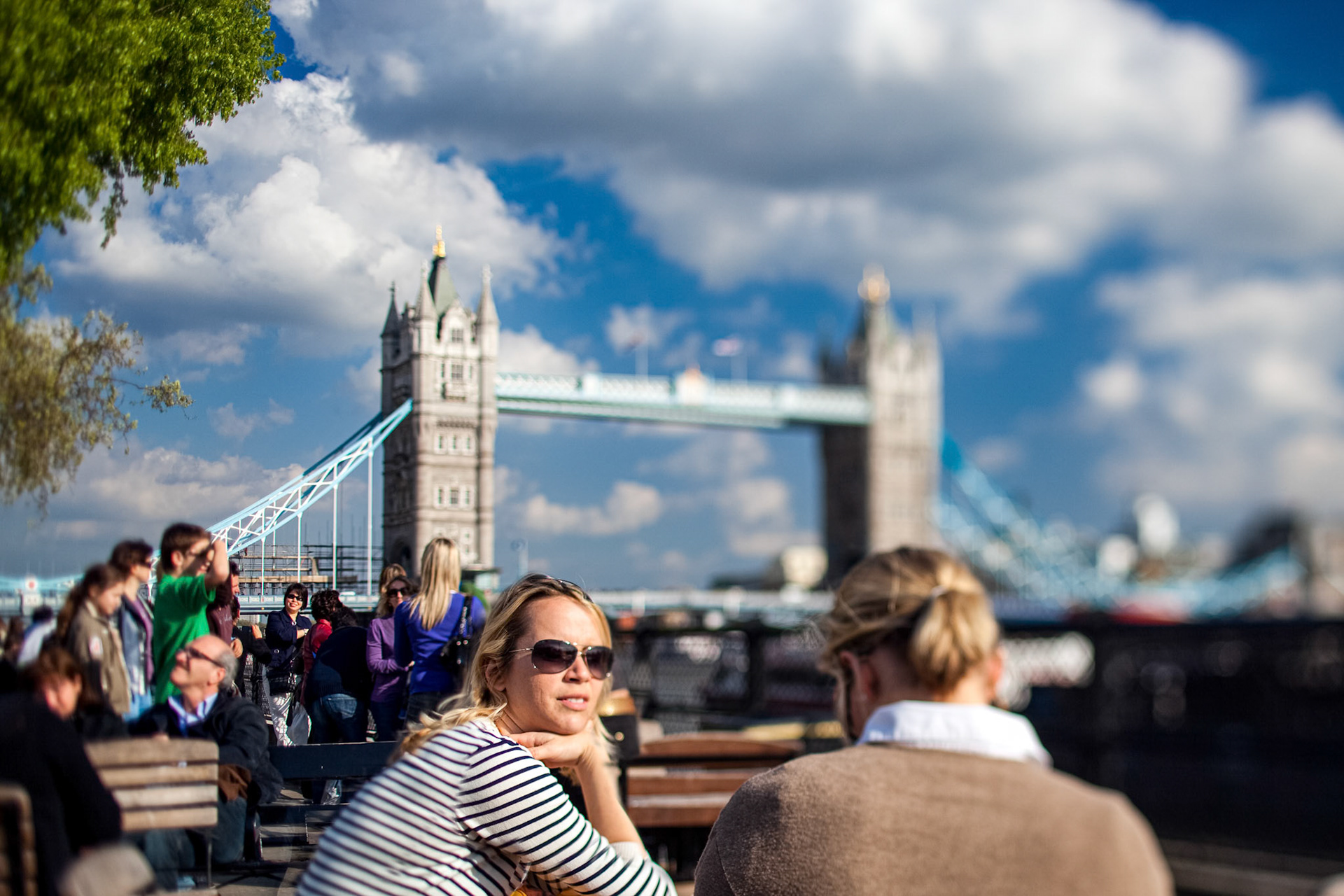 Visitors relax on the north bank of the Thames river while Tower Bridge stands tall in the background. Clouds drift above in a busy London scene.