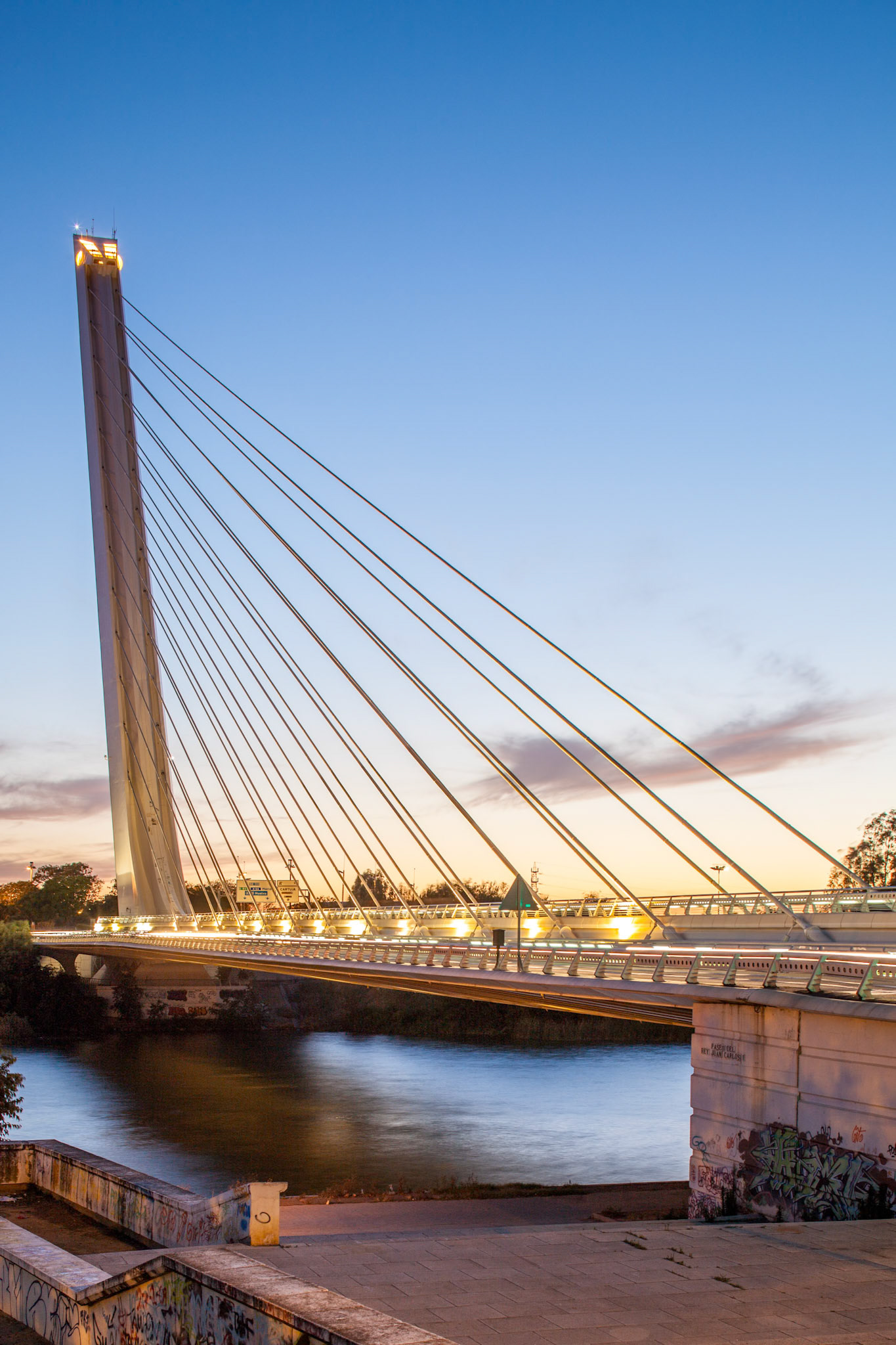 Alamillo Bridge is illuminated at dusk, reflecting on the calm waters of the Guadalquivir River in Seville, creating a serene atmosphere.