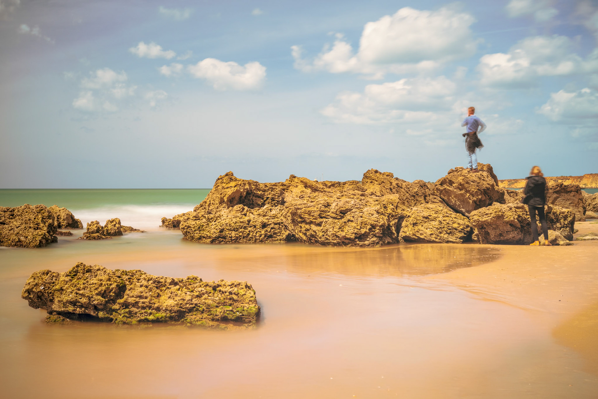 Couple walks along Roqueo beach, exploring mossy rocks and wet sand under a moody sky. Long exposure reveals shimmering textures in the coastal scene.