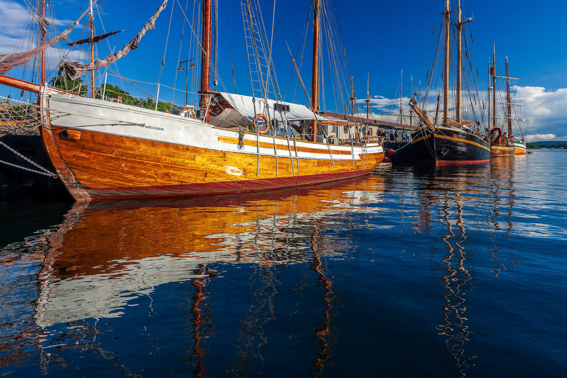 Boats and sailing ships rest peacefully at Aker Brygge dock in Oslo, reflecting warm sunlight on serene water.