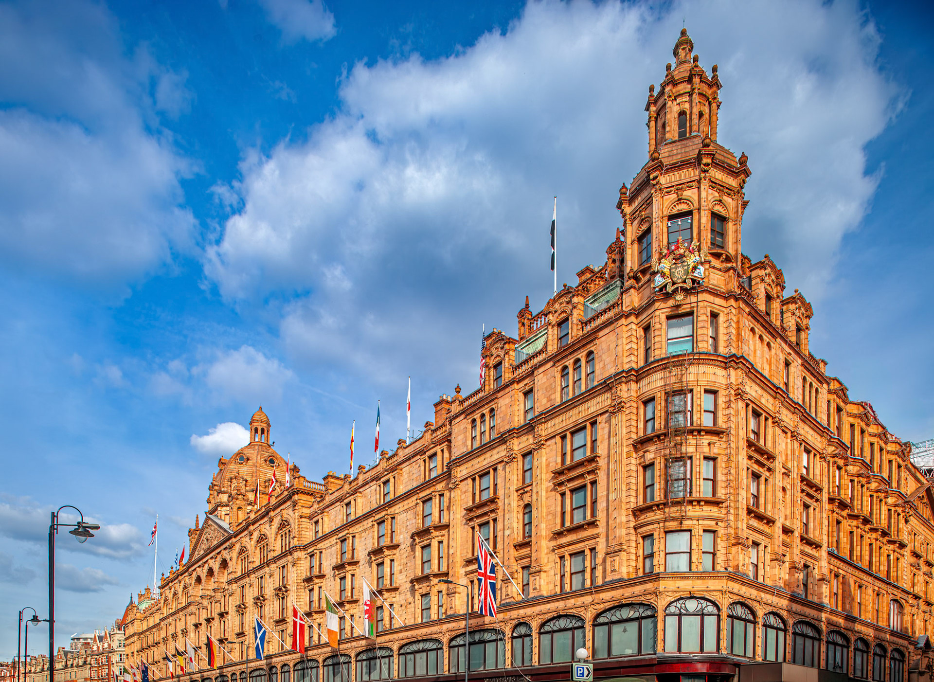 The photo shows the Harrods building, a famous store in London, on Brompton Road. Clouds are in the sky, and flags are displayed on the building.