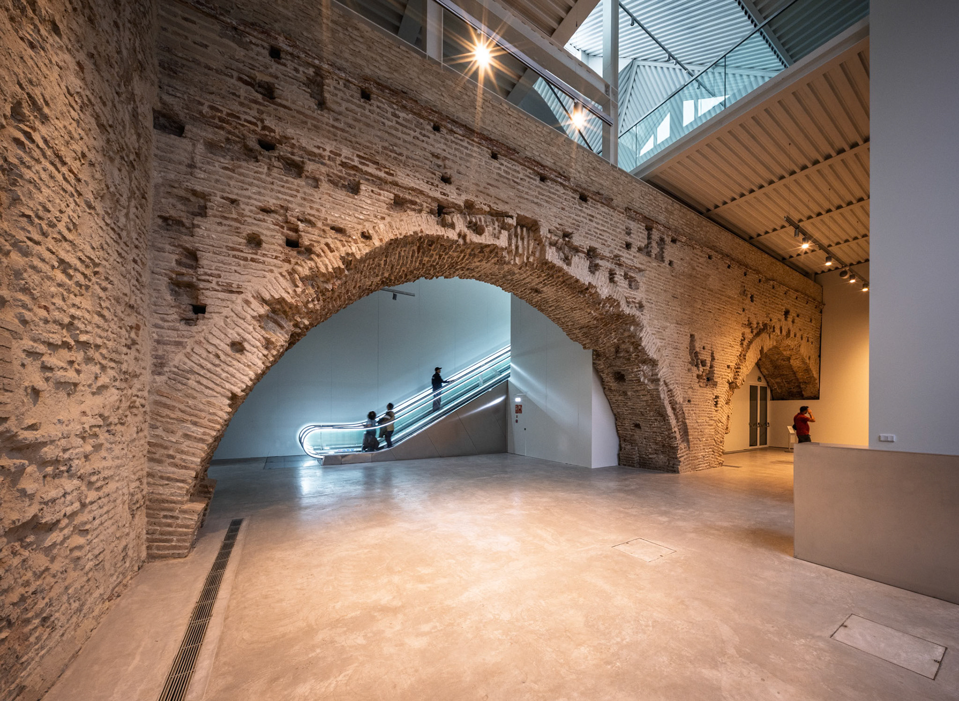 Visitors ascend an illuminated escalator through a medieval brick arch in Seville's Atarazanas, linking the ground to the first-floor exhibitions.