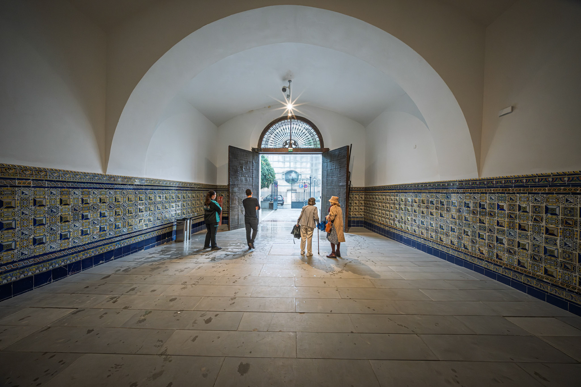 People gather in the vaulted entrance of Seville’s Atarazanas, admiring patterned tiles and open iron doors under a stunning transom.