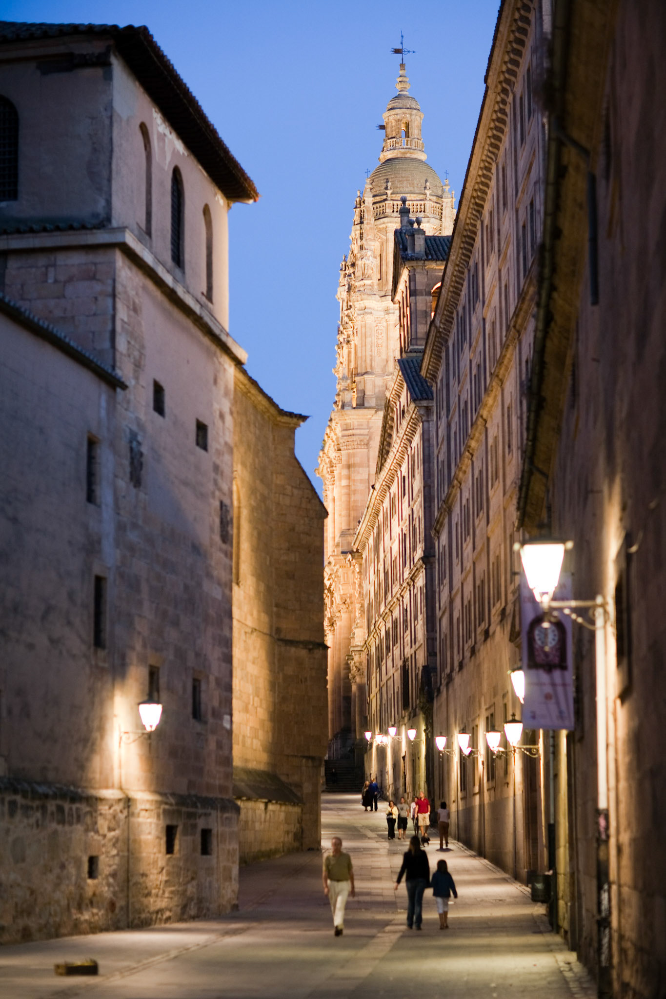 Visitors stroll down Calle de la Compañía, admiring historic architecture as the University Pontifica and Clerecía towers light up dusk.