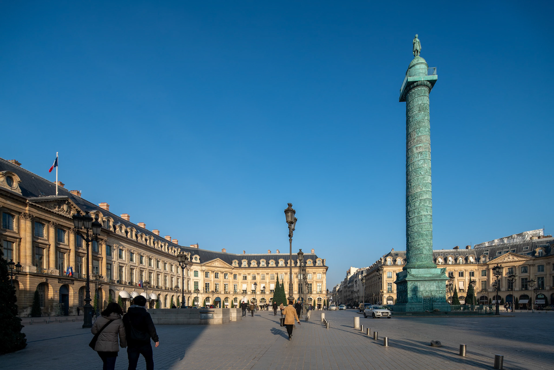 Pedestrians walking near the Vendome Column under a clear blue sky.