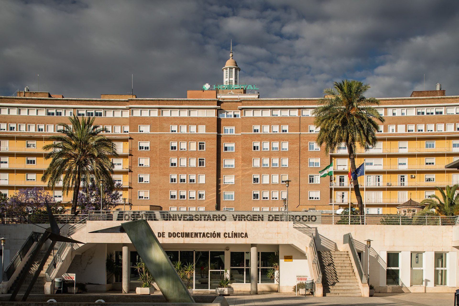 Virgen del Rocío Hospital in Seville, inaugurated in 1955, features modern architecture with palm trees and a cloudy sky.