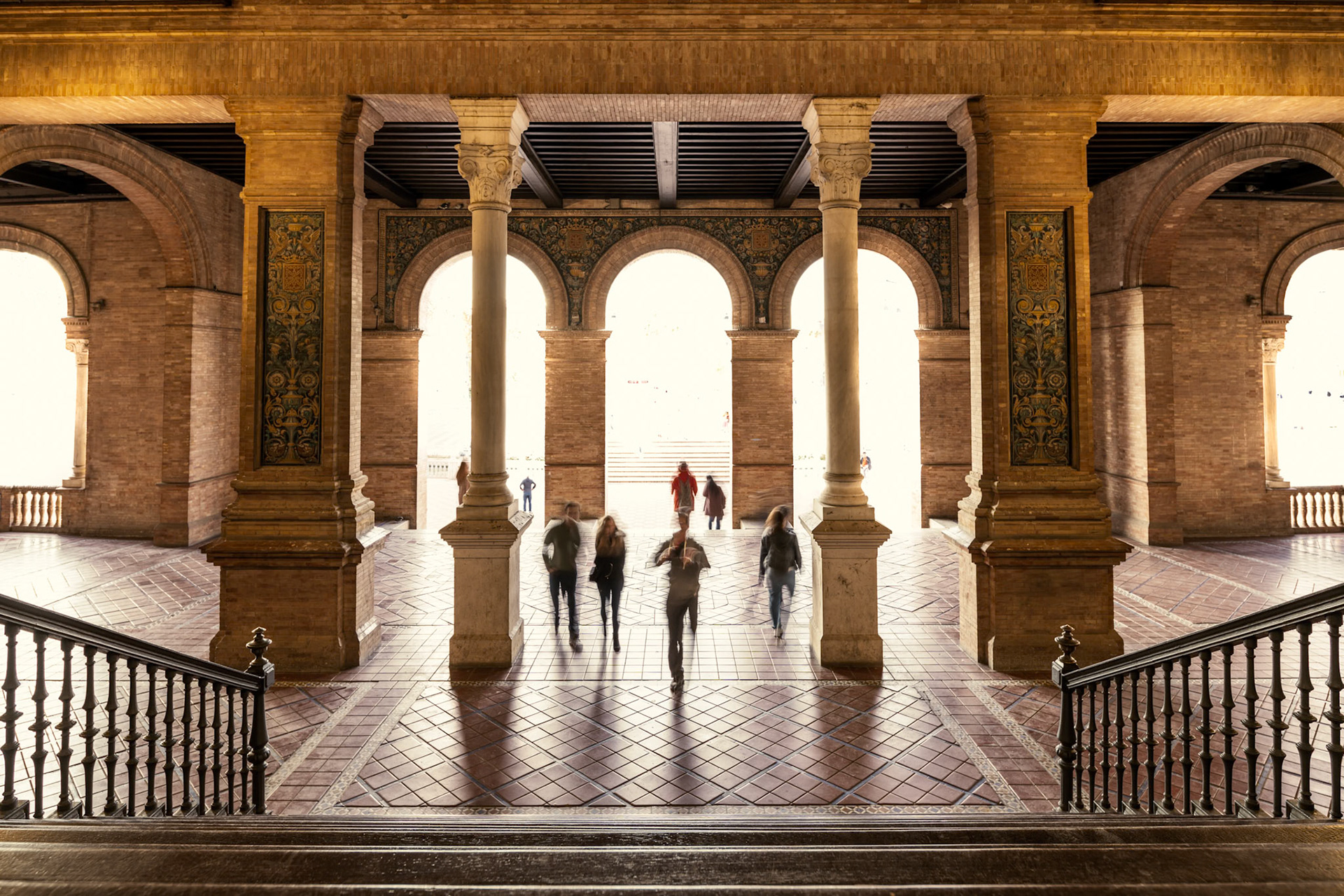 Blurred figures glide through the arched gallery of Plaza de España, merging motion with the stunning tiled architecture of Seville.