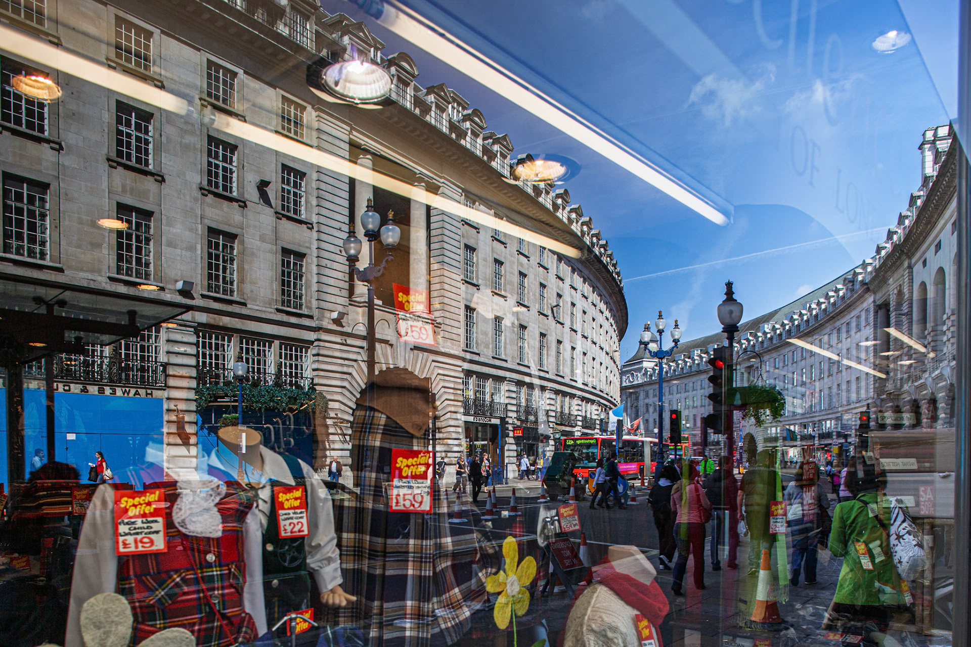 A busy shopping area can be seen through the reflections on a shop window in Regent Street. People walk by as traffic moves in the background.