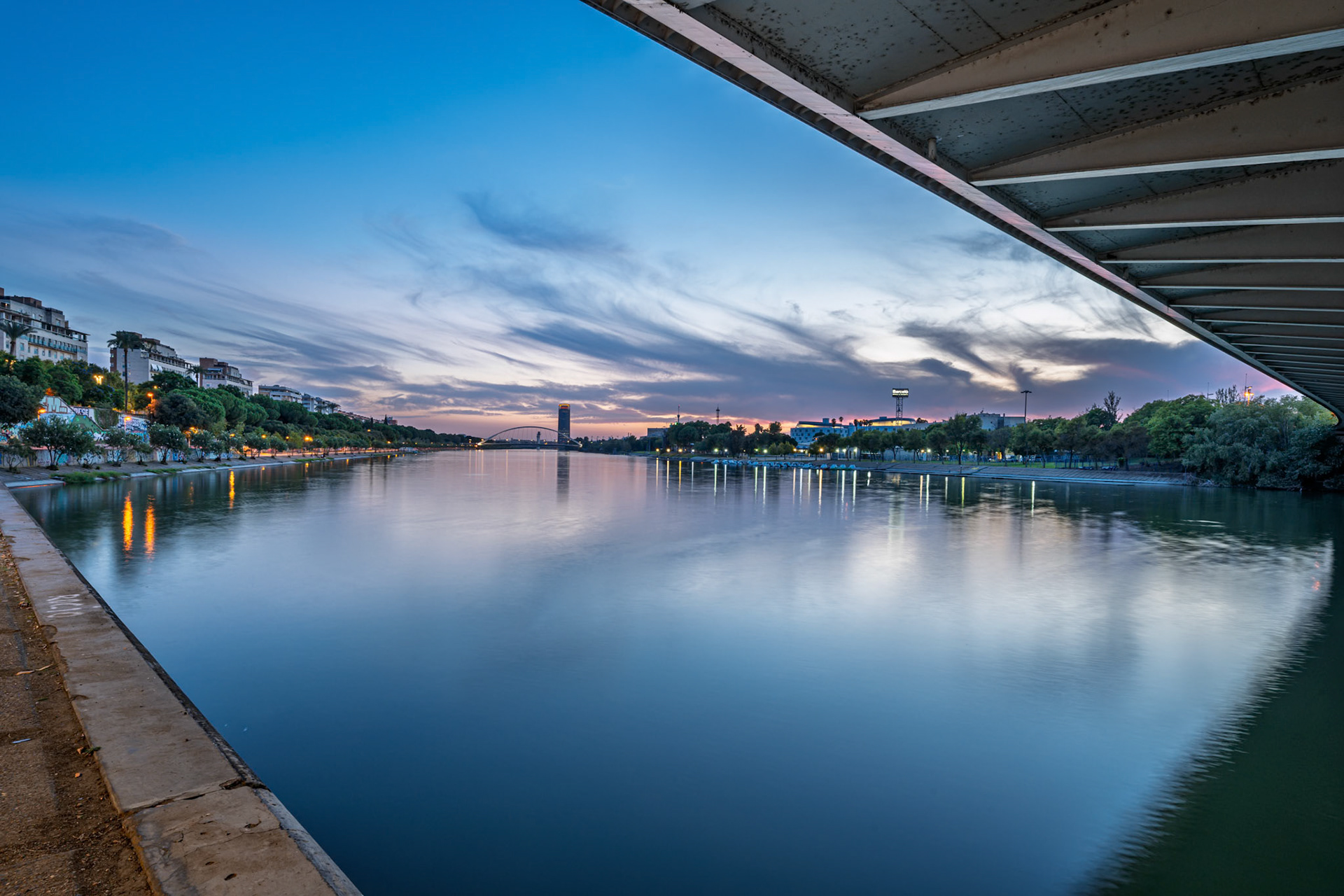 Sunset casts beautiful colors over Guadalquivir River, highlighting La Cartuja Island and Torre Sevilla while reflecting on calm waters.