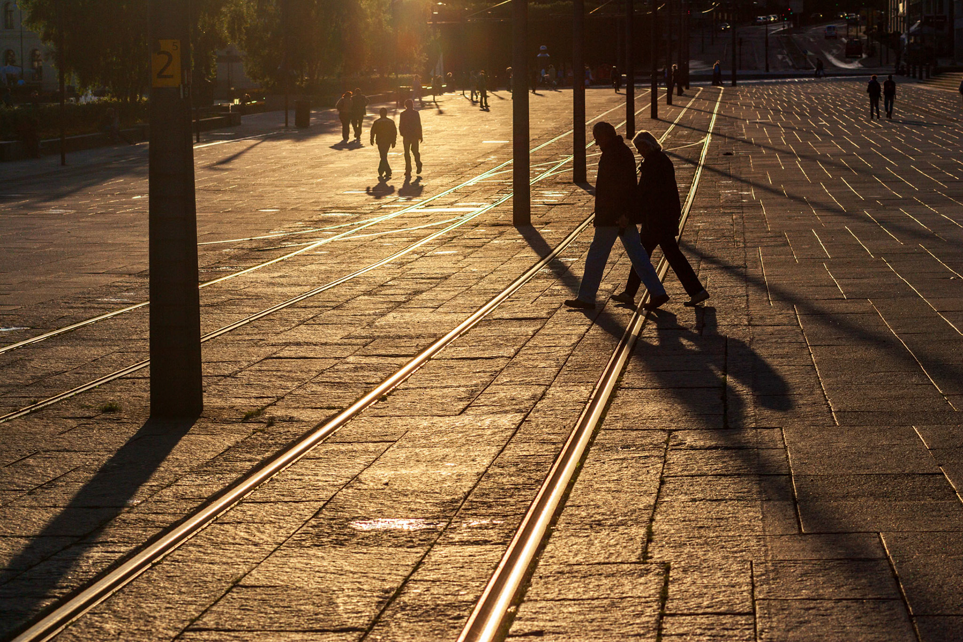 At sunset in Oslo, two walkers cross the gleaming tram lines while others stroll along the sunlit path.