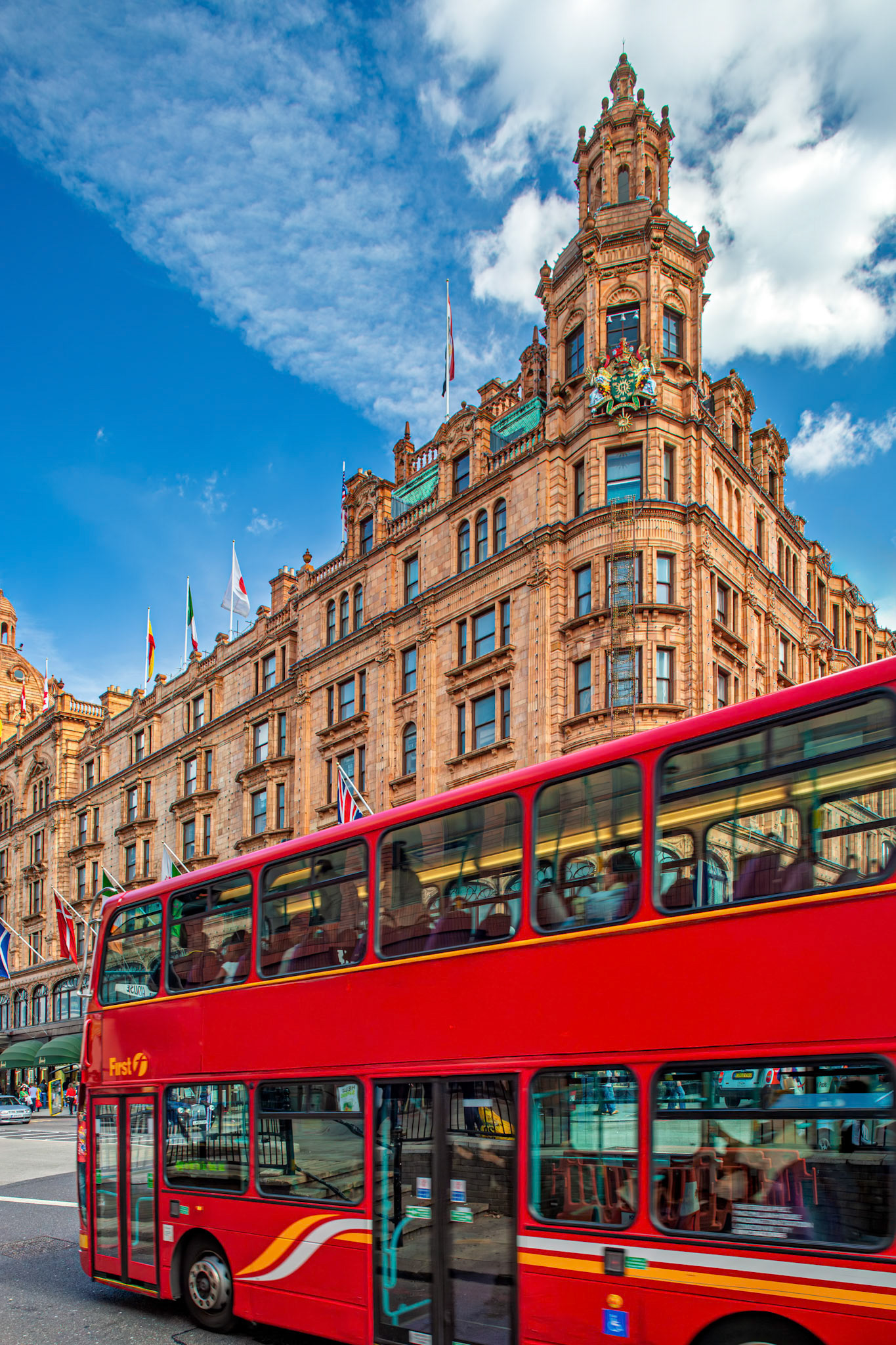 A red double-decker bus moves along Brompton Road. Harrods stands tall in the background with flags waving under a clear blue sky.