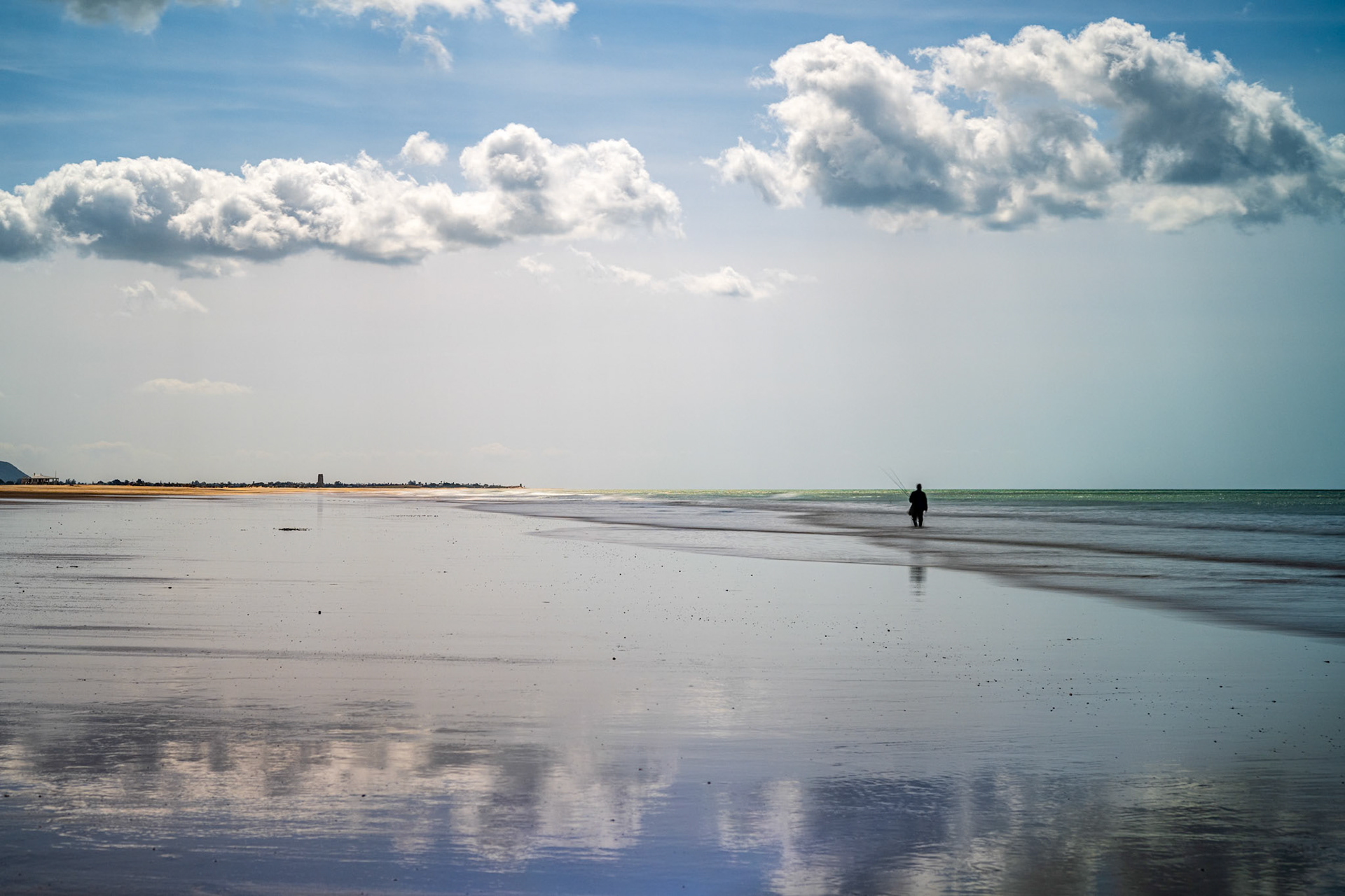 A lone fisherman stands on the wet sand of Conil's winter beach, casting his line into the calm Atlantic waters, creating a peaceful seascape.