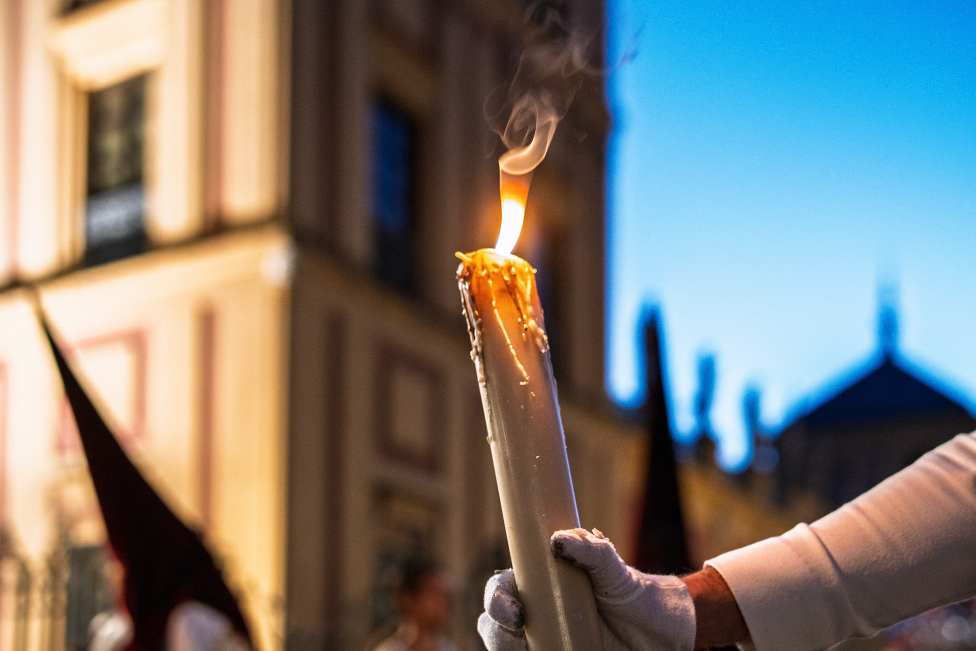 Warm candlelight glows as a penitent holds a candle in Seville, capturing the solemnity of Holy Week at twilight.