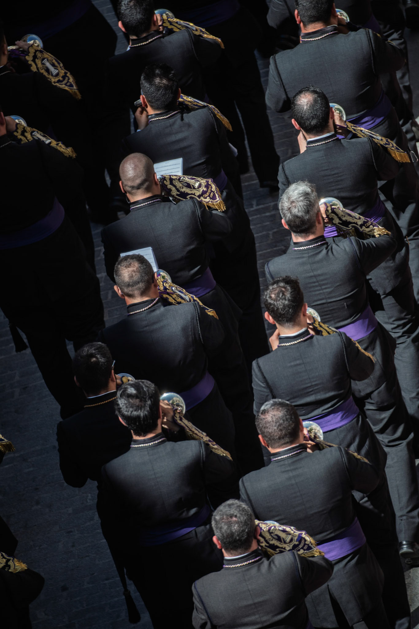 Participants in dark attire march in a solemn procession, playing brass instruments as part of Holy Week celebrations in Seville.