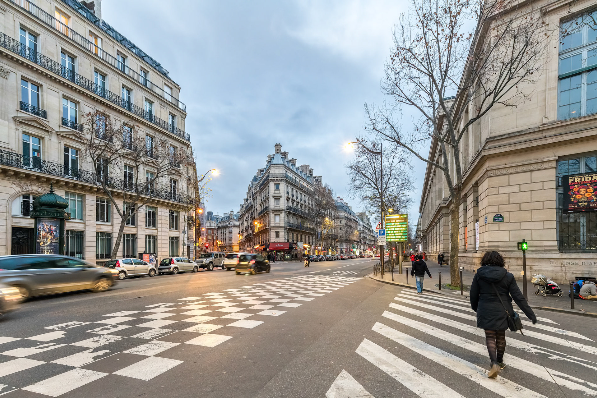 Pedestrians cross at twilight on the bustling streets of Paris with classic architecture lining the boulevard.
