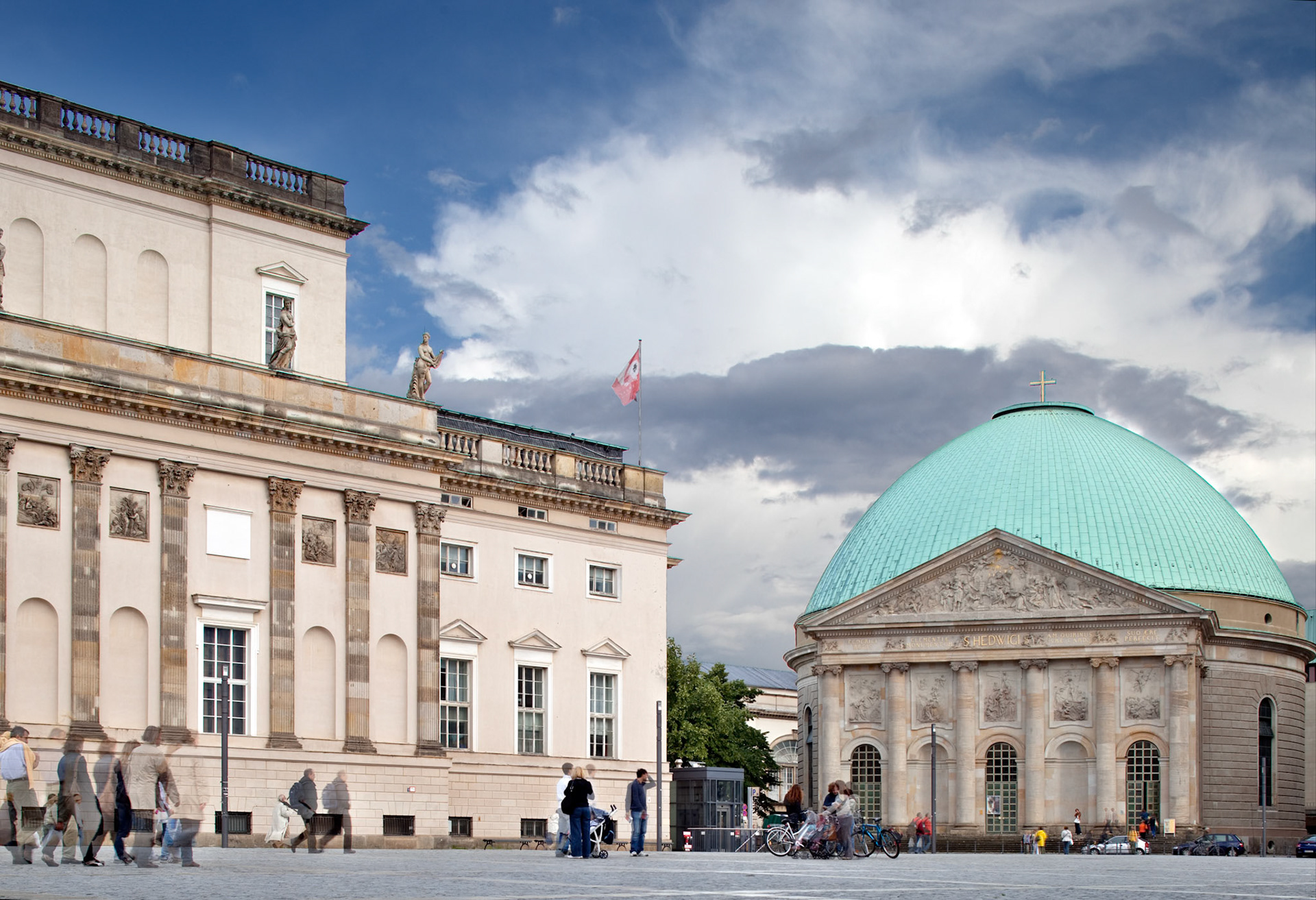 People stroll through Bebelplatz, showcasing the Staatsoper and Saint Hedwig Cathedral against a cloudy sky.