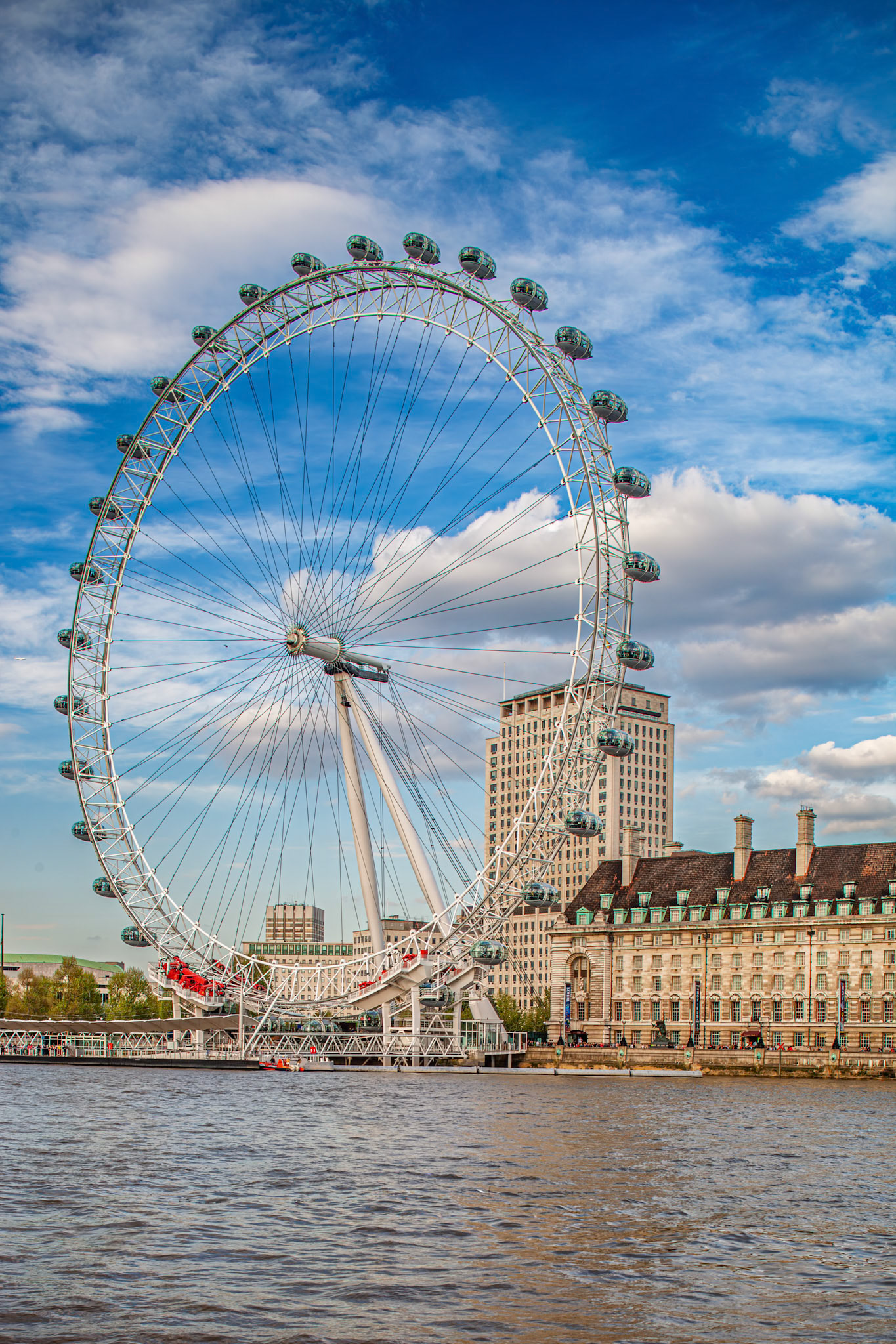 London Eye stands tall on the banks of River Thames with clouds above and buildings nearby. Visitors enjoy sightseeing from the capsules.