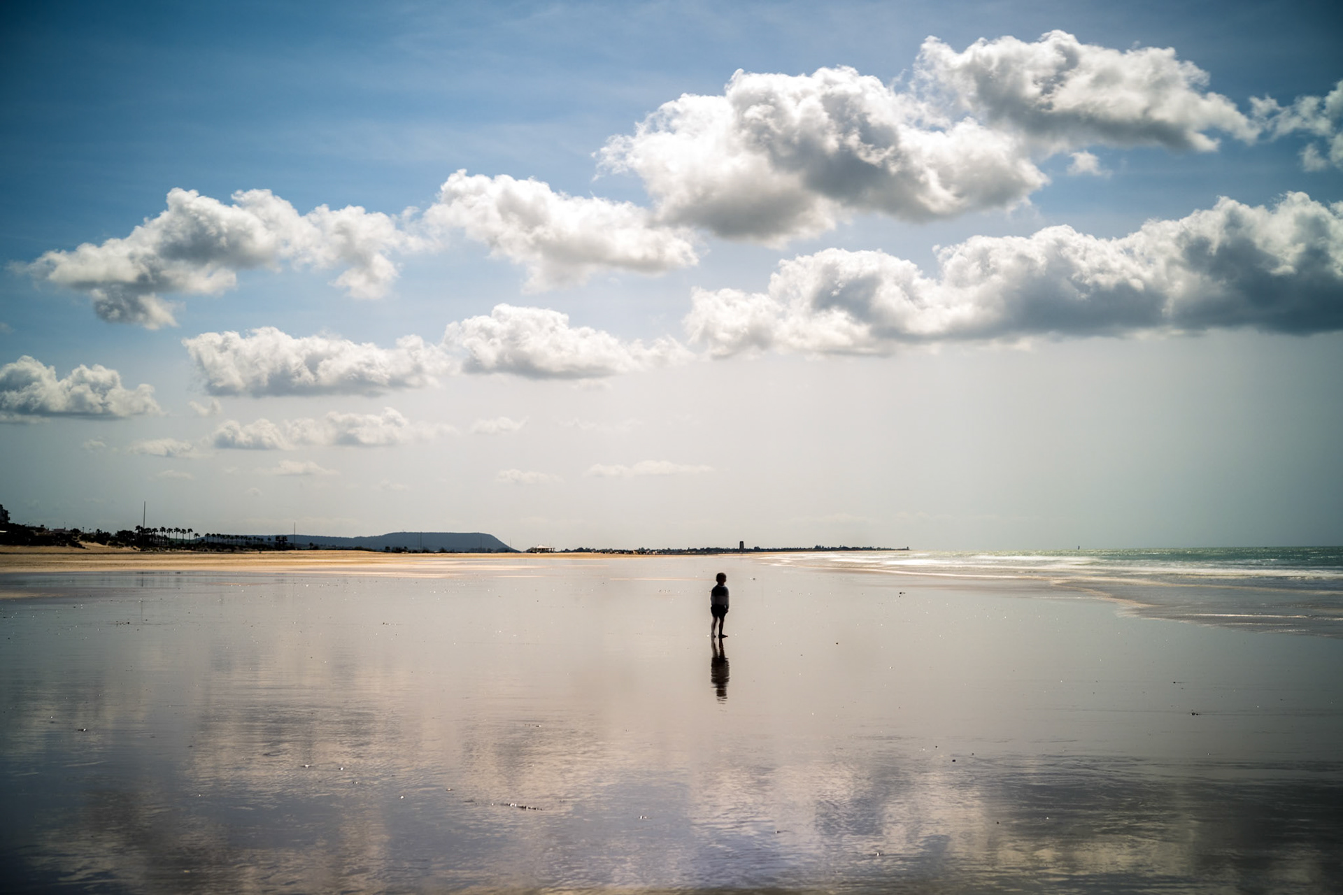 A child stands alone on the winter beach in Conil de la Frontera, reflected in wet sand under a soft sky.