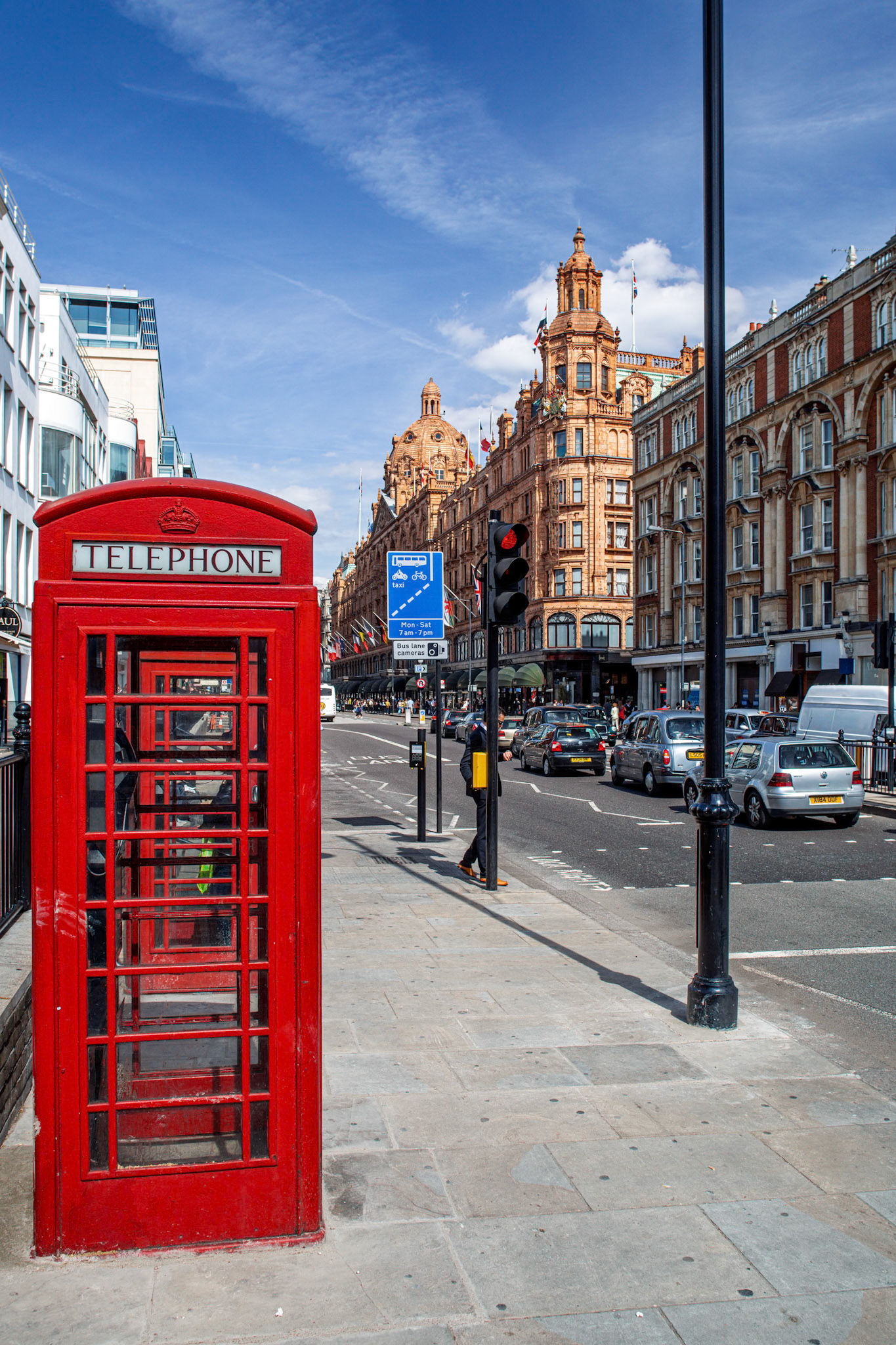 Two red telephone boxes stand on Brompton Road with the Harrods building visible in the background. Traffic flows along the busy street in London.