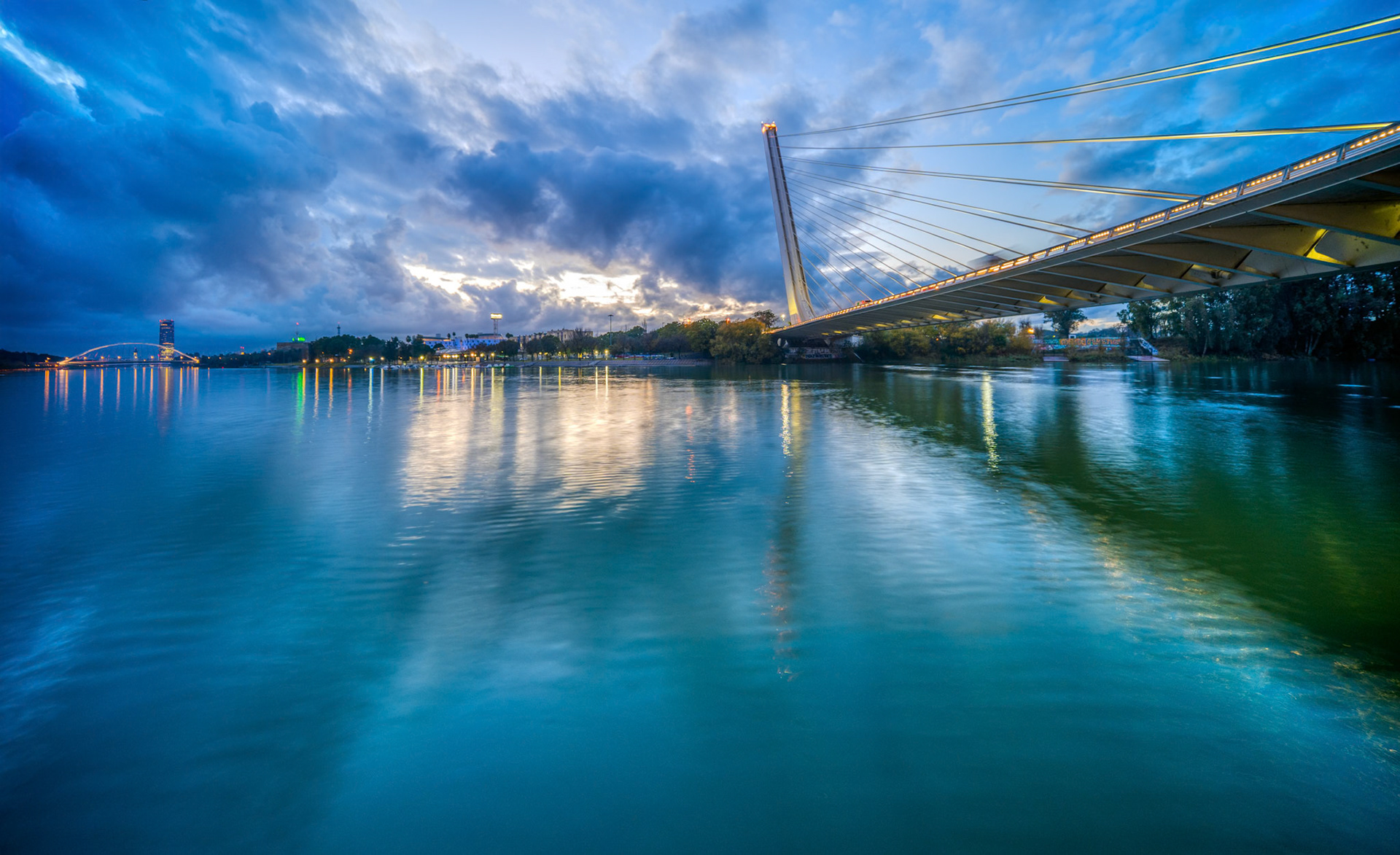 Wide-angle view of Seville's riverfront at dusk, showcasing Alamillo Bridge and Barqueta Bridge with reflections on the Guadalquivir River.