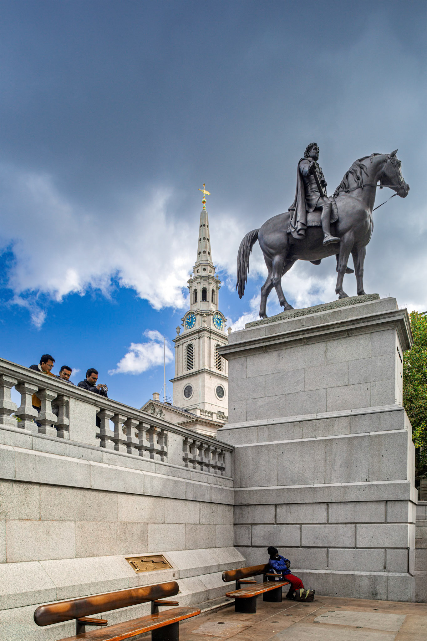 Visitors stand near the George IV statue at Trafalgar Square, London. St Martin in the Fields bell tower is visible in the background under a cloudy sky.