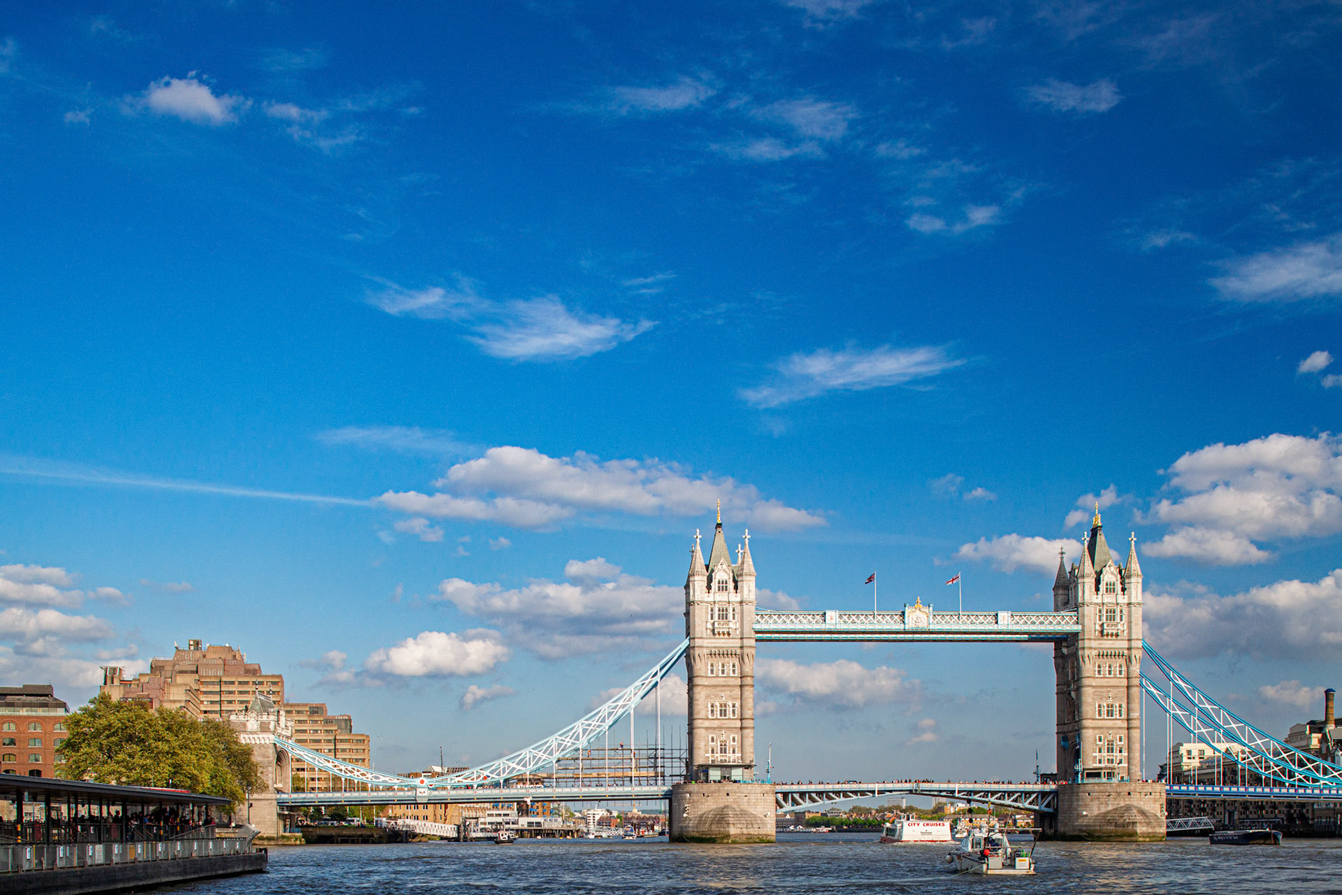 People enjoy a sunny day by the Thames River while observing Tower Bridge. Boats travel beneath the bridge with blue skies in the background.