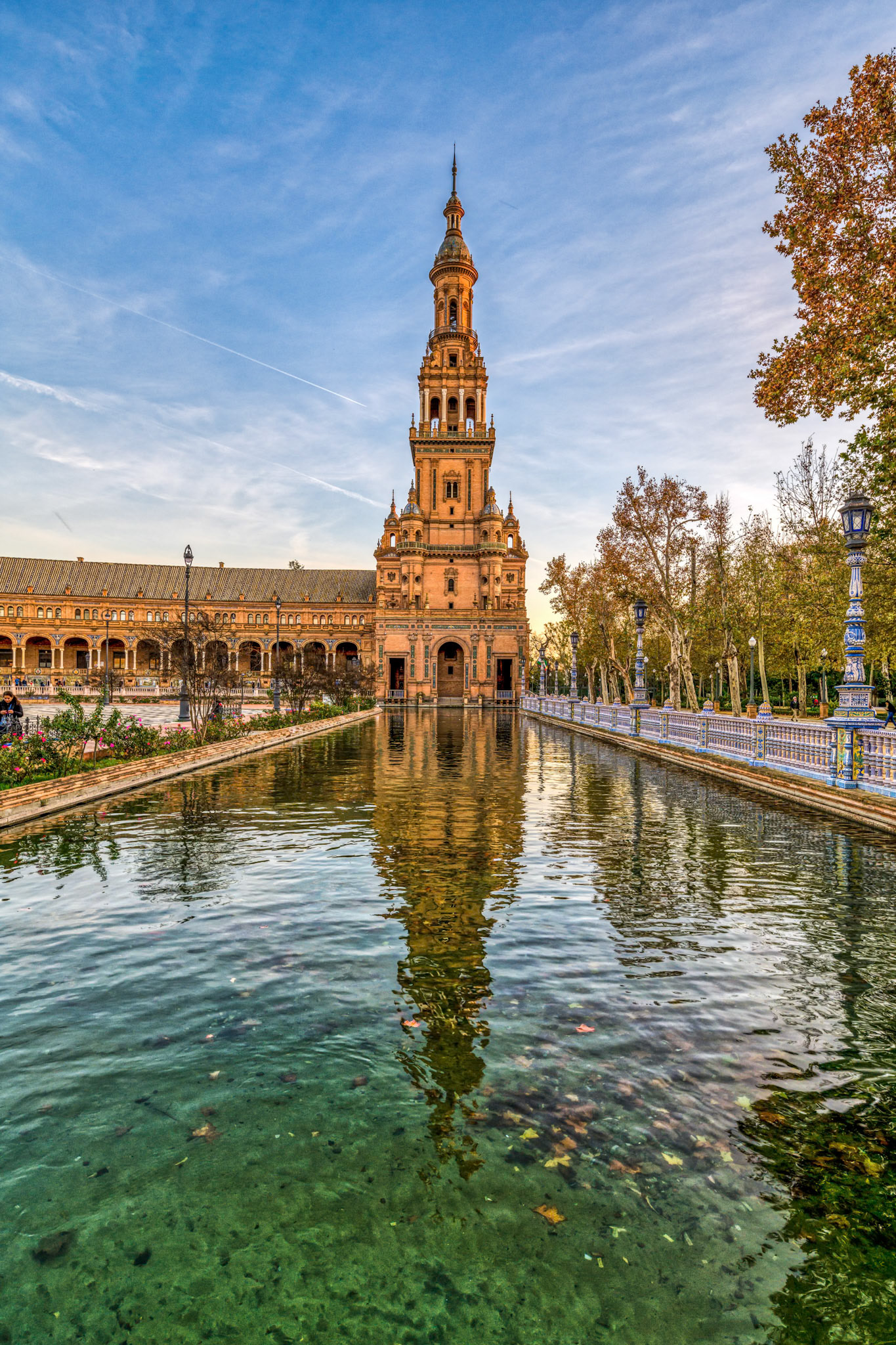 The south tower of Seville’s Plaza de España rises majestically at sunset, perfectly mirroring in the calm waters below.