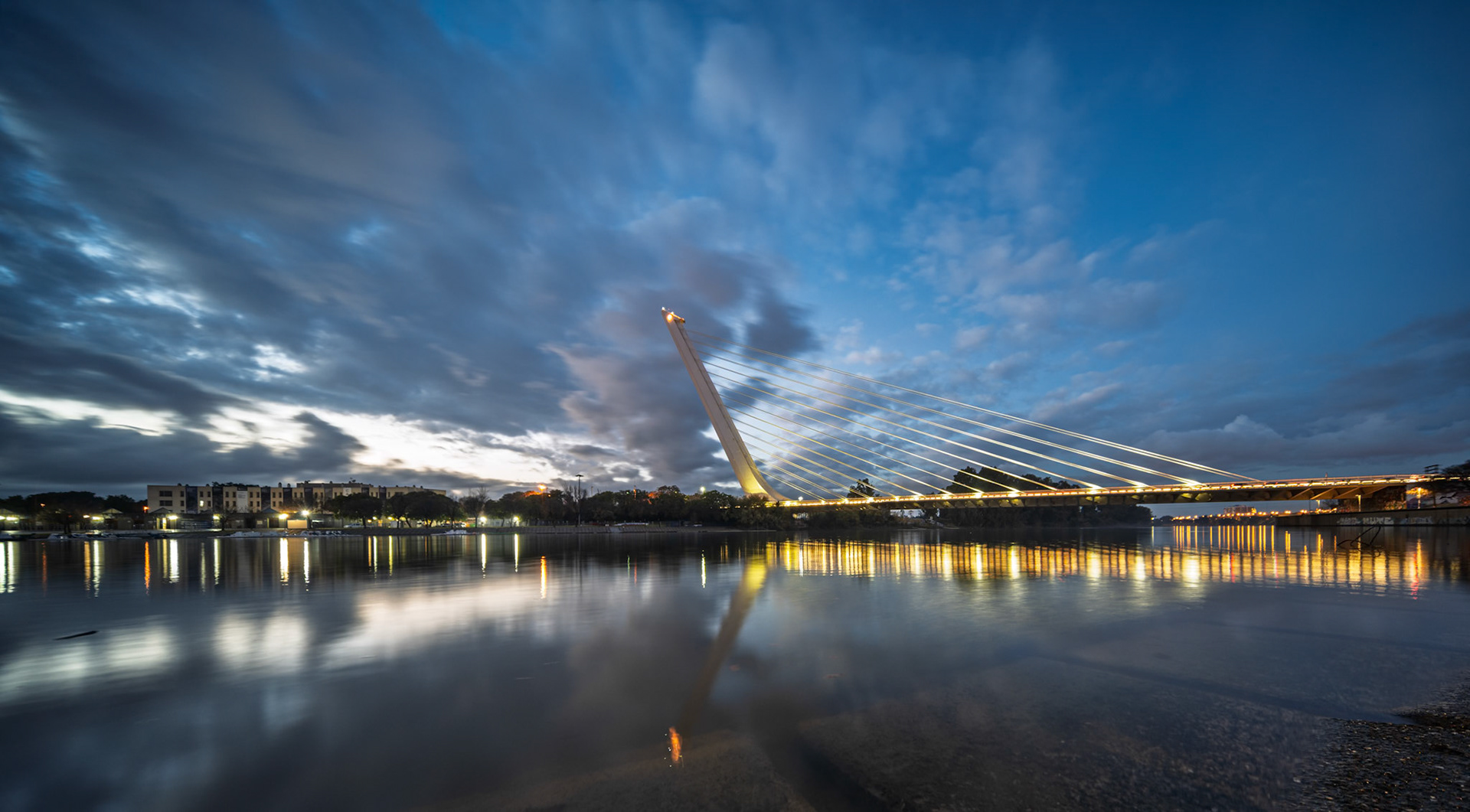 Alamillo Bridge in Seville is illuminated at dusk, showcasing long exposure effects on the calm water and vibrant sky filled with clouds.