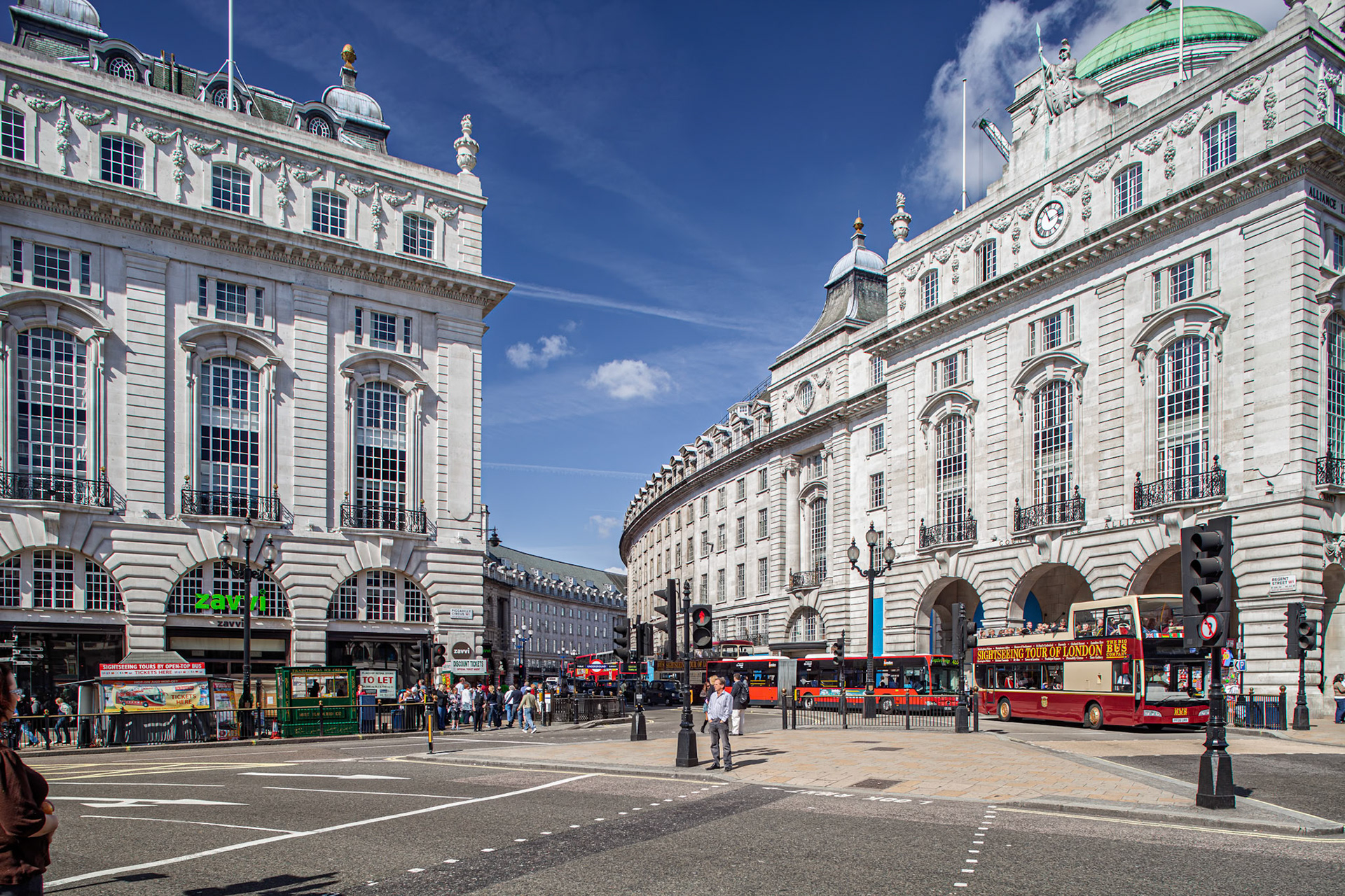 Many people gather at the busy intersection of Regent Street and Piccadilly Circus. Red buses and shops are visible in the background.